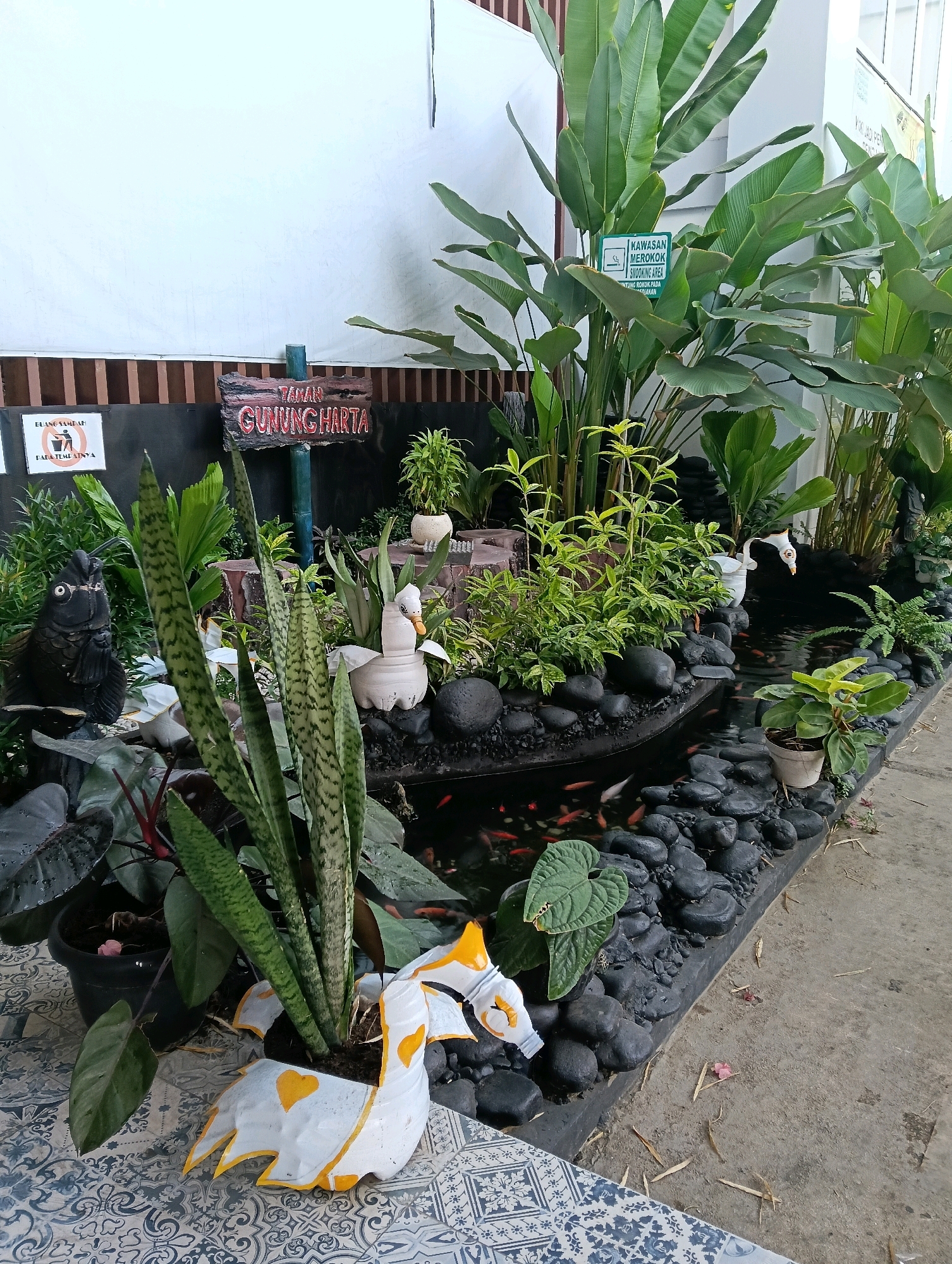 The image depicts an outdoor garden area, likely a small pond and plant display. In the background, a white wall or banner is visible, with a wooden fence and some signage. One sign, in red lettering on a rustic wooden plaque, reads "TAMAN GUNUNG HARTA," indicating it's the "Gunung Harta Garden." Another green sign, a "No Smoking" sign, is also present amongst the foliage.
The foreground features a variety of plants, including tall, broad-leafed greenery and smaller, spiky succulents like snake plants. The pond itself is lined with dark, smooth stones and contains numerous small, orange goldfish swimming in dark water. Decorative elements include a black, fish-shaped statue with glasses, and two white swan-shaped decorations made from repurposed plastic bottles, one of which is painted with yellow accents and a yellow heart. A distinctive white and yellow planter, also possibly made from recycled materials in the shape of a stylized animal or creature, sits in the lower left foreground. The ground around the pond is a mix of dark stones and concrete, with some scattered dry leaves and a small pink petal. The overall scene suggests a decorative garden or public space, possibly a rest area or part of a larger attraction. Based on the signage, the location is identified as Taman Gunung Harta, within the city of Magelang, Indonesia. There are no people visible in the image.