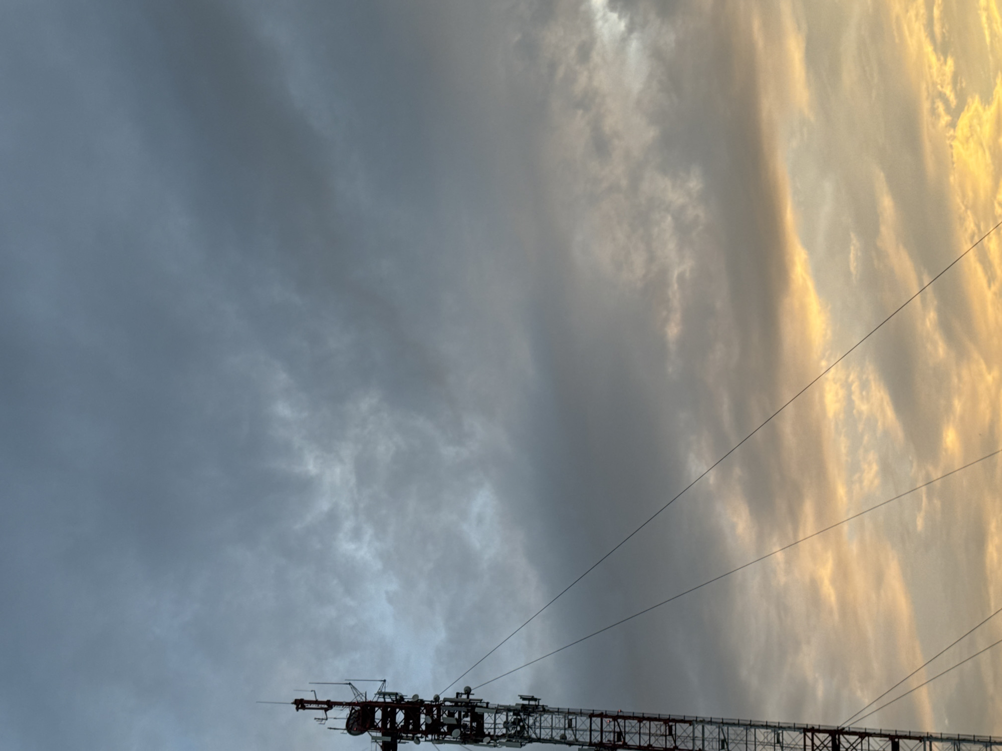 An outdoor, low-angle, vertical shot captures a cell tower against a dramatic sky. The tower, painted red and white, dominates the left side of the frame, with its complex structure of antennas and equipment visible. Several guy wires extend diagonally from the tower, disappearing out of frame. The sky is filled with heavy, grey clouds, but towards the bottom right, the clouds are illuminated by the golden hues of sunset or sunrise, creating a striking contrast. The scene appears to be set in or near the city of Zaokskiy, Russia, given the context of the location. There are no people or any other discernable objects in the immediate vicinity.