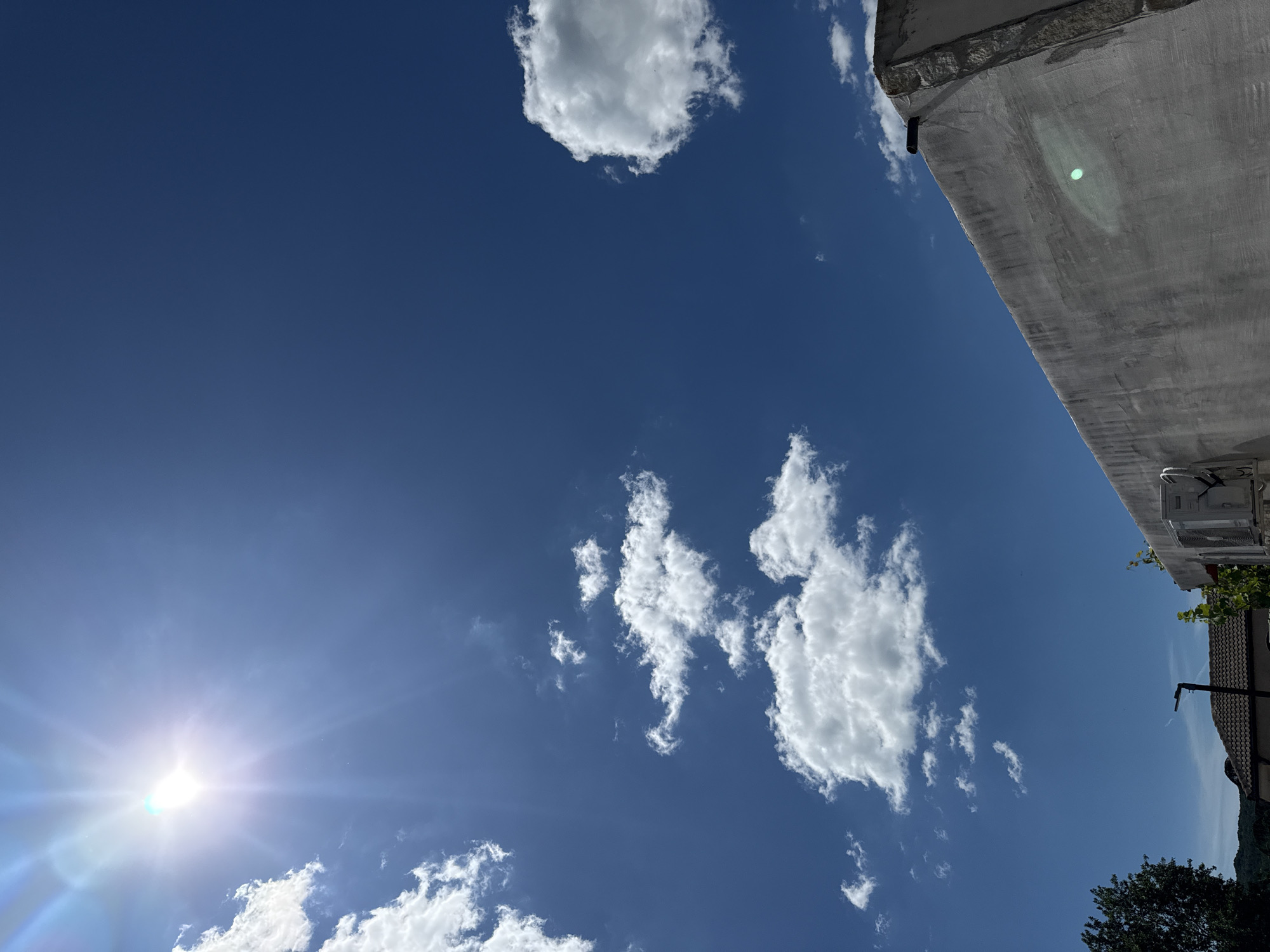 A bright, sunny day is depicted with a clear blue sky. The sun is high in the sky and appears to be casting bright rays. There are a few white, fluffy clouds scattered across the sky. In the foreground, a gray, concrete wall runs diagonally across the lower right portion of the frame. The wall appears to be part of a building, and it has some texture and a few cracks. An air conditioning unit is mounted on the wall. In the lower left corner, a portion of a rooftop with dark shingles is visible, along with a lamppost. A few trees and distant hills are also discernible in the background. The overall scene suggests an outdoor environment on a warm day.