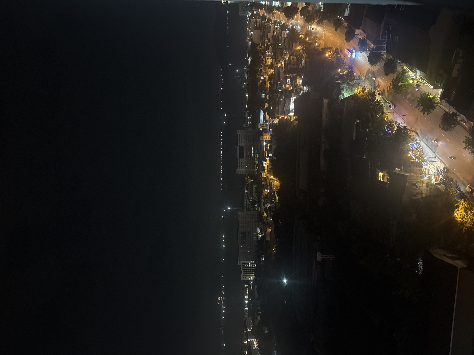 The image depicts a nocturnal cityscape in Nha Trang, Vietnam, viewed from a high vantage point. The scene is dominated by the dark expanse of the sky, with scattered lights of the city below and the glimmering surface of the ocean in the distance.
In the foreground, the top of a dark building is visible, with some illuminated windows. Below this, the cityscape unfolds with a network of streets and buildings. Numerous streetlights cast a warm, yellow glow on the asphalt, illuminating the paths of vehicles. Several cars and a bus are visible on the road, their headlights and taillights creating streaks of light. Pedestrians are present on the sidewalks, though their details are indistinct due to the low light.
Further into the city, larger buildings, possibly hotels or apartment complexes, are illuminated with a brighter, white light. The silhouette of trees is noticeable, interspersed with the urban structures.
The middle ground shows a body of water, likely the ocean, stretching towards the horizon. Scattered lights on the water suggest boats or distant coastal structures. The horizon is hazy, with faint lights visible, indicating a wider coastal area.
The overall impression is one of a vibrant city coming alive at night, with the ocean serving as a backdrop to the urban development. The image captures the characteristic nighttime ambiance of a coastal city, with the interplay of artificial lights and the natural darkness.