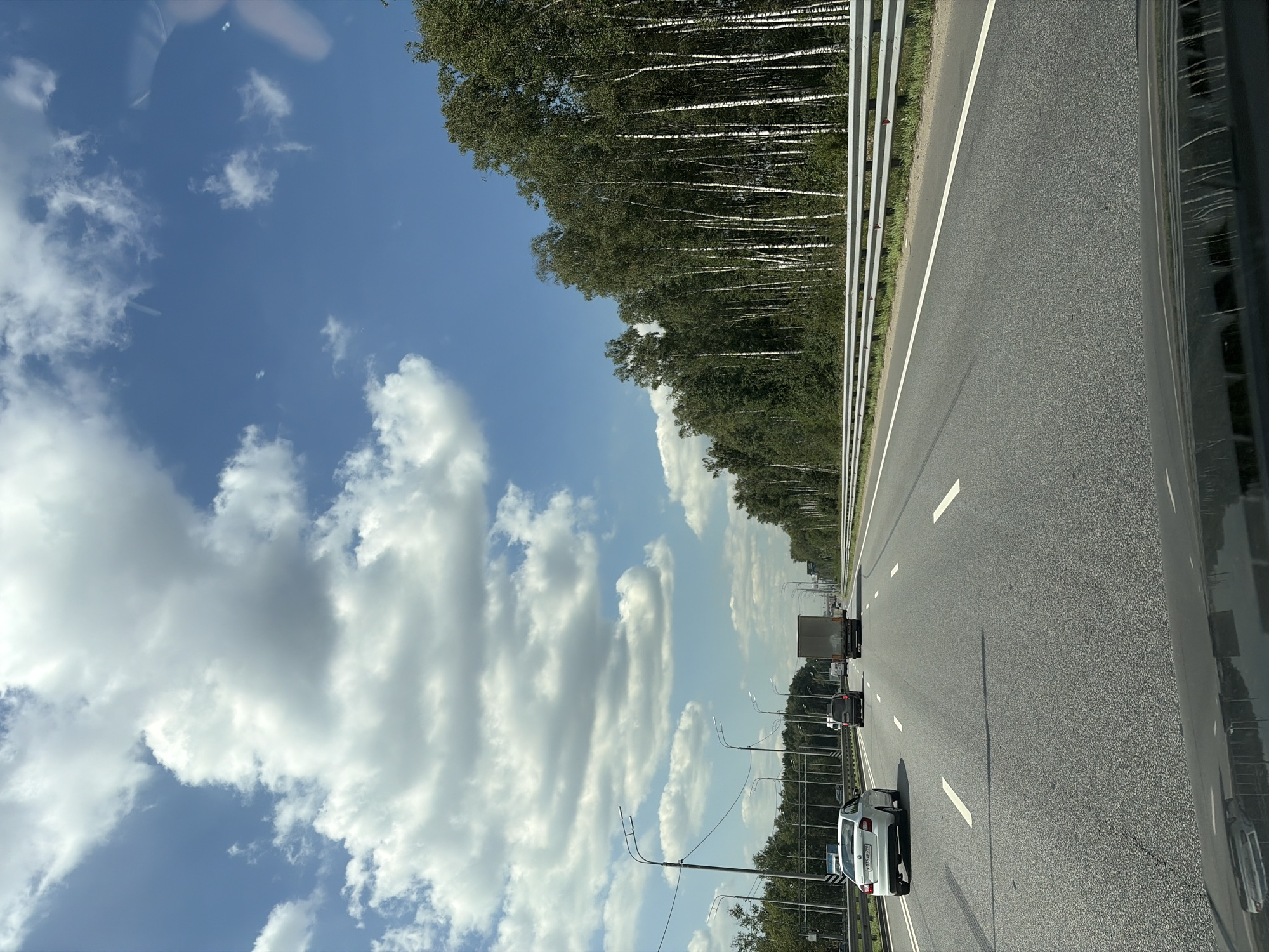 This is a vertically oriented, eye-level, outdoor, daytime, medium shot of a highway in Russia, possibly near Эфир Снт. The perspective is from inside a moving vehicle, looking towards the right side of the road. A bright blue sky with scattered white clouds fills the upper left portion of the frame, with trees lining the left side of the highway. The highway itself is asphalt, marked with white lane lines, and appears to have at least two lanes in the direction of travel.

Several vehicles are visible on the highway, including a white car in the foreground, a dark SUV, and a truck further ahead. Lampposts are spaced along the right side of the highway. The trees appear to be a mix of deciduous trees, possibly birches, given their white trunks. The overall mood is peaceful and suggests a journey or travel. There are no people clearly visible as subjects in the frame. The scene is bright, indicating it is likely midday. The license plate of the white car in the foreground is partially visible and appears to read "X736AP 777".