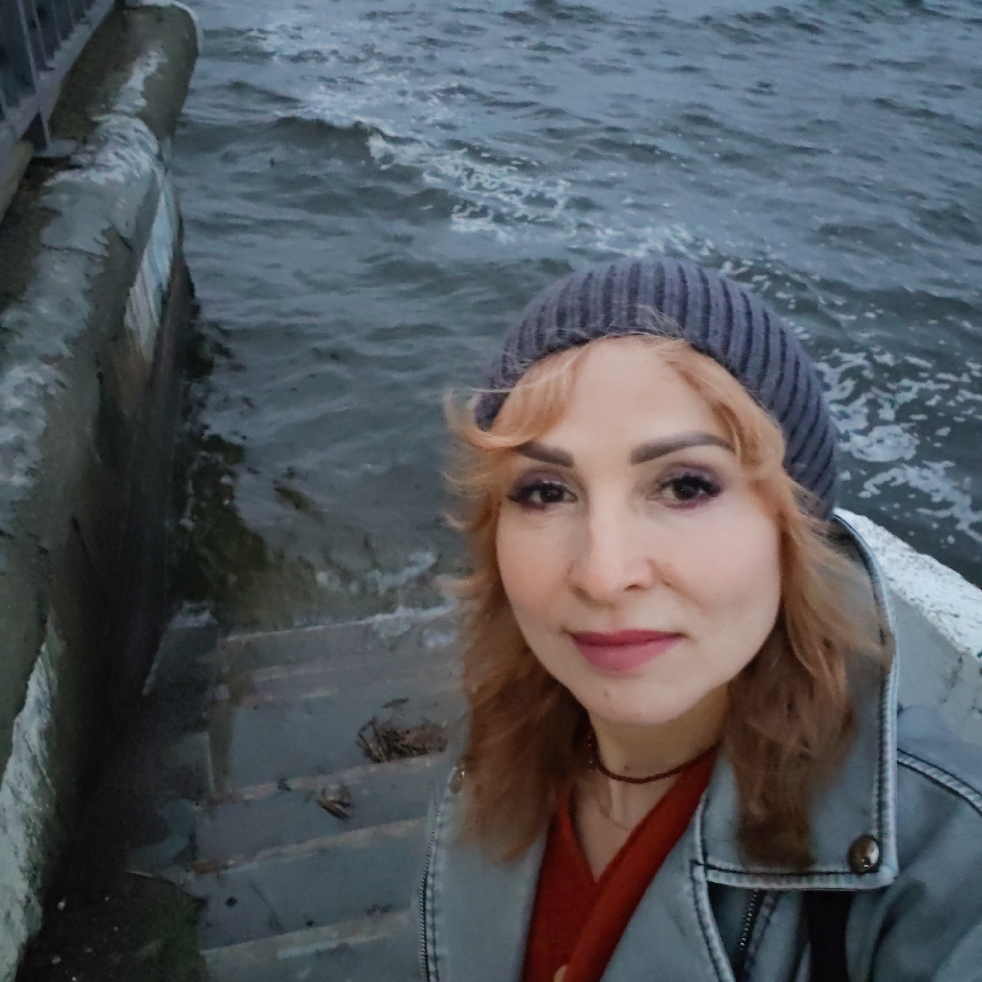 A woman stands on concrete steps near choppy water. She wears a dark knitted cap, a red top, and a light-colored denim jacket. Her hair is a light auburn color. The steps lead down to the water, with a concrete barrier visible on the left side. The location is Chaikovsky, Russia.