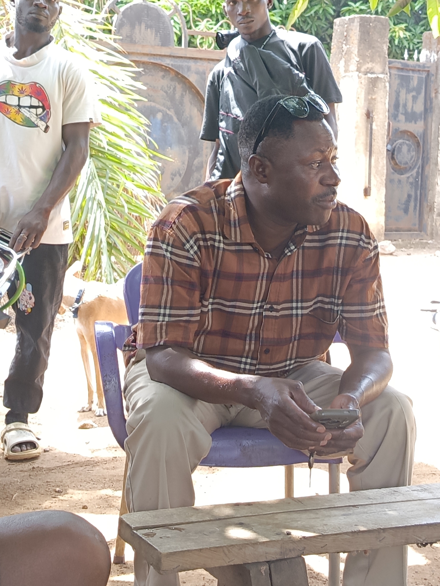 Outside, in Akwanga, Nigeria, a man sits on a purple plastic chair, holding a cell phone, with his head turned to his right, wearing a plaid shirt and khaki pants. A wooden bench is in front of him. Another man stands to his left, wearing a white t-shirt with a lip print with a marijuana leaf, dark pants and white sandals. A third man is standing in the background behind the seated man, wearing a dark t-shirt. A dog is standing behind the seated man. A metal gate is behind the men. Green foliage is also visible in the background.