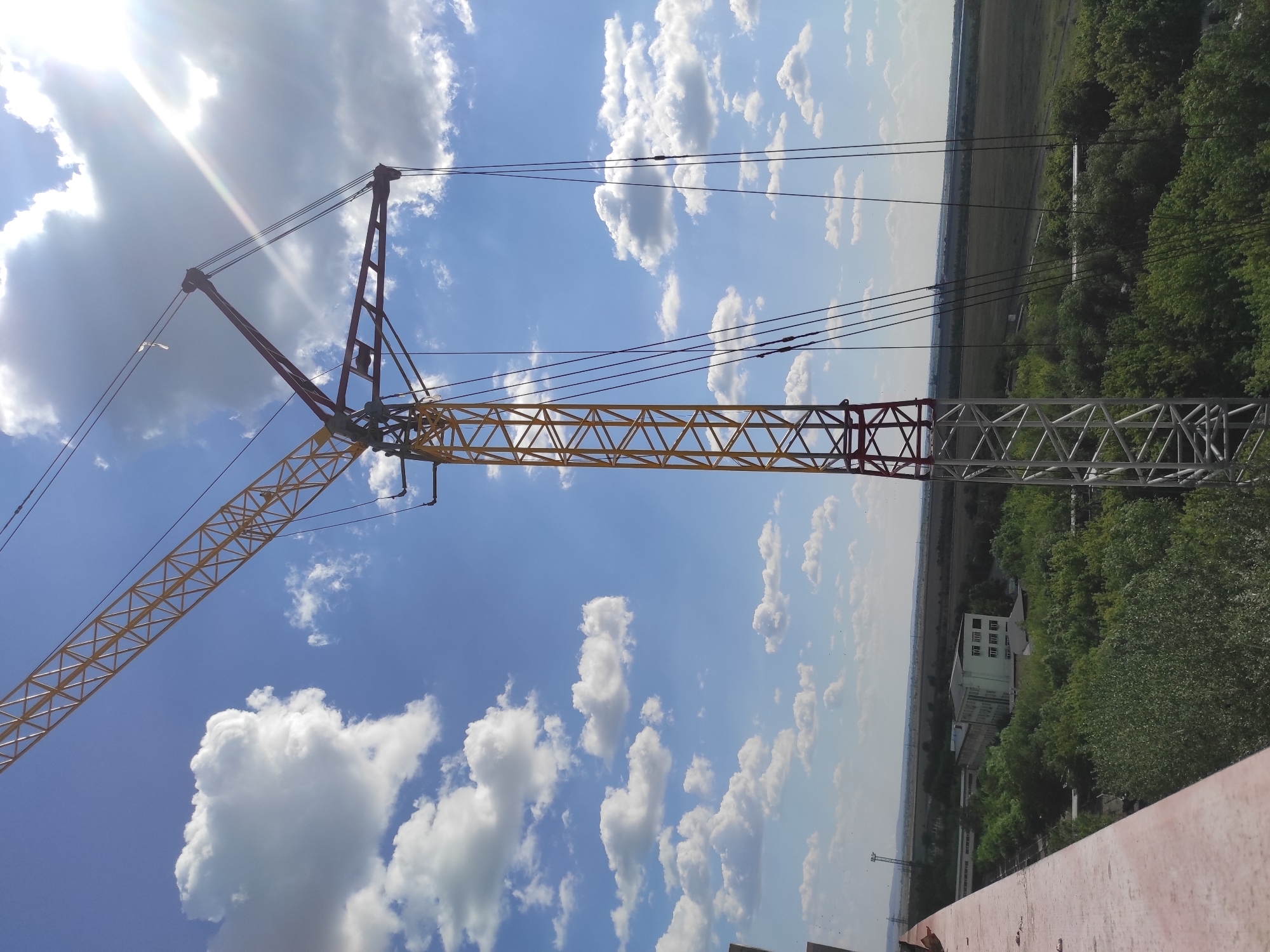 A construction crane, with a yellow lattice boom and red and white sections of its vertical support structure, is prominently displayed against a bright blue sky with scattered white cumulus clouds. The sun is visible in the upper right corner, casting a strong glare and lens flare. The crane's cables hang down, some partially obscured by the structure. In the background, a green landscape stretches to the horizon, dotted with trees and a long, rectangular building with multiple windows. The foreground features a portion of a red-brown, weathered structure, possibly a rooftop or balcony, on the left side of the frame. The image appears to have been taken from a high vantage point, looking out over a semi-urban or industrial area. The specific location within Kyzyl, Kazakhstan, cannot be determined from the visual information provided.