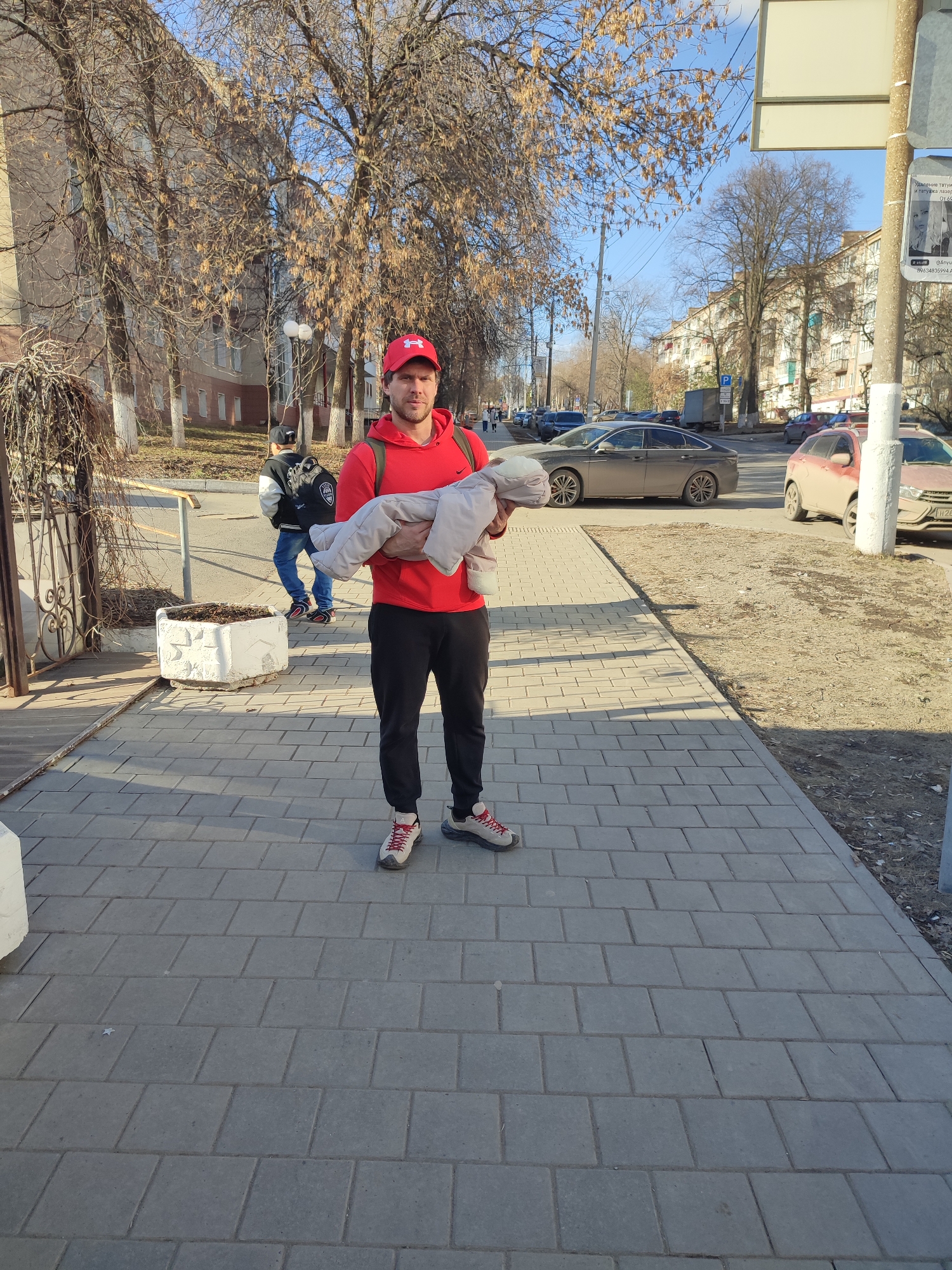 A man stands on a gray brick sidewalk in Izhevsk, Russia. He wears a red Under Armour hat, red hooded sweatshirt, black athletic pants, and sneakers. The man holds a bundle wrapped in light gray fabric. Behind him to the left, a person wearing a black and white jacket and jeans walks away on the sidewalk. A potted plant sits adjacent to the sidewalk on the left. In the background, parked cars line the street. Buildings and bare trees are also visible in the background. A street sign is posted on a pole to the right.