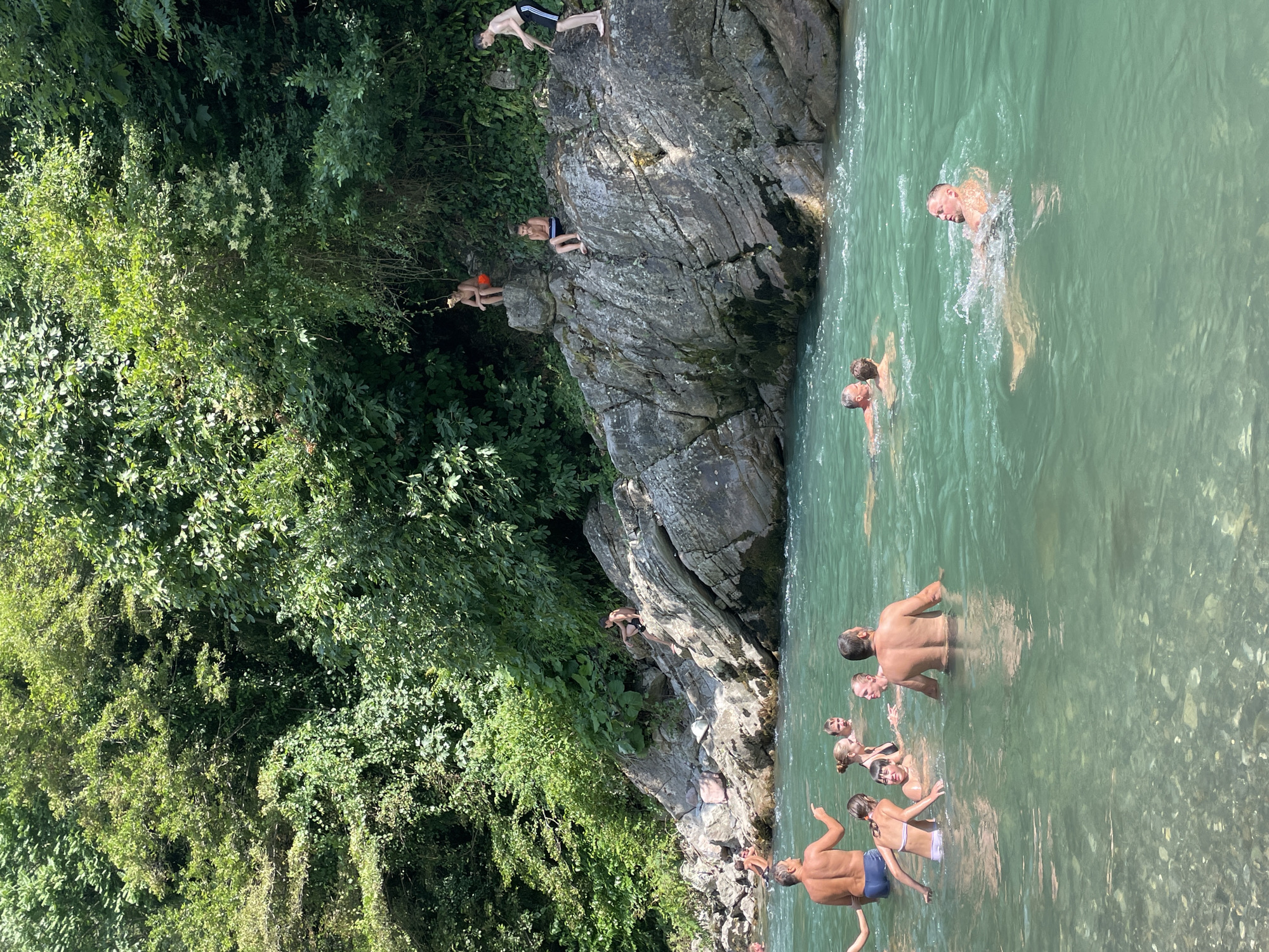 This media file shows a group of people swimming and jumping into a clear, turquoise river on a sunny day. The scene is set in a natural environment with lush green foliage and large rocks surrounding the river. Several individuals are visible on the rocks, with some appearing to be preparing to jump into the water. Others are already in the water, swimming and enjoying the refreshing river.
The time of day appears to be midday, with bright sunlight casting shadows and highlighting the textures of the rocks and the clarity of the water. The weather is clearly warm and sunny, conducive to outdoor recreational activities. The location seems to be a popular spot for swimming and cliff jumping, indicated by the presence of multiple people and the natural rock formations that lend themselves to such activities. The general atmosphere appears relaxed and joyful, with people engaged in leisurely water activities. No visible text is present in the image.