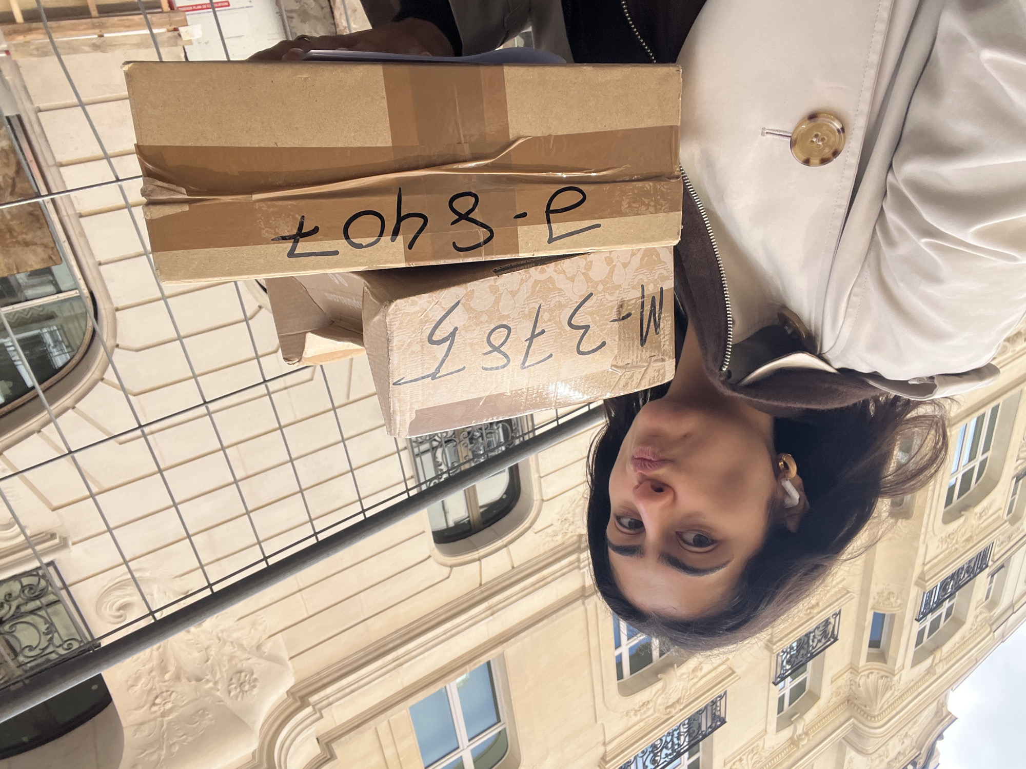 A medium shot captures a person holding two brown cardboard boxes outdoors, with Parisian architecture visible in the background. The person, a woman with dark hair and brown eyes, is positioned so that her face is partially visible. She wears an earbud in one ear and a beige jacket with a brown collar and a large brown button. The boxes are stacked, with the top box displaying the handwritten text "P-8407" and the bottom box reading "M-3787." The background features a building with ornate facades, iron balconies, and numerous windows. The building's architectural style is indicative of Paris, France. A metal barrier with vertical and horizontal bars is visible in the foreground, partially obscuring the building.