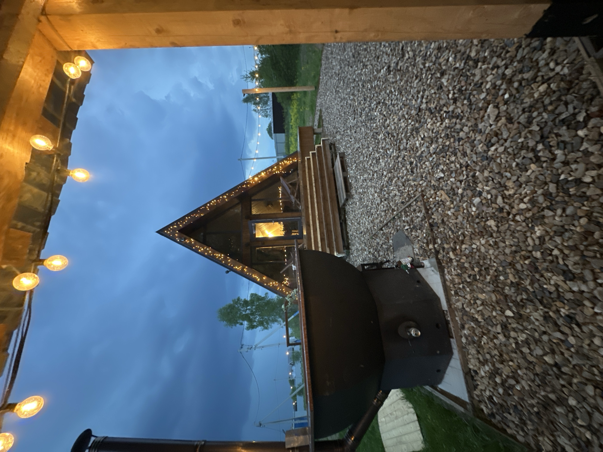 The image shows a view from a wooden structure, possibly a porch or entryway, looking towards a small A-frame cabin at twilight. The cabin is decorated with string lights along its roofline. The surrounding ground is covered with gravel. A large, dark metal object, potentially a grill or smoker, is situated on a concrete slab next to the gravel area. In the distance, there are other buildings and trees. String lights are visible in the background. The location is Малеевка, Russia.