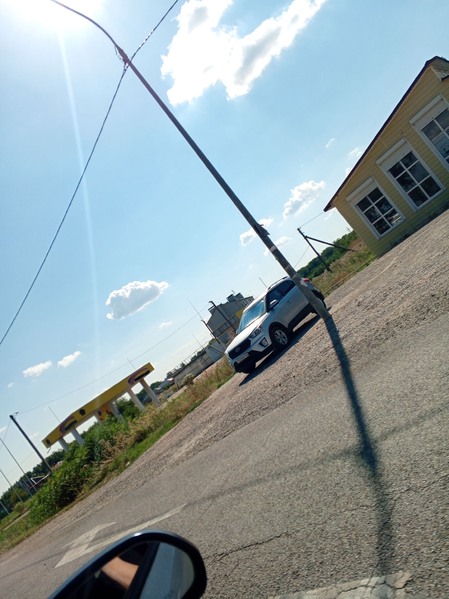 The image depicts a sunny outdoor scene, likely in a rural or suburban area. A silver SUV is parked on a gravel surface next to a paved road. In the background, there is a yellow building with white-trimmed windows, and a structure that appears to be a gas station canopy with yellow beams. A utility pole with wires runs diagonally across the frame, casting a shadow on the road. The sky is blue with scattered white clouds, and the sun is creating a lens flare in the upper left corner. The perspective suggests the photo was taken from inside another vehicle, with a portion of a side-view mirror visible in the bottom left corner.