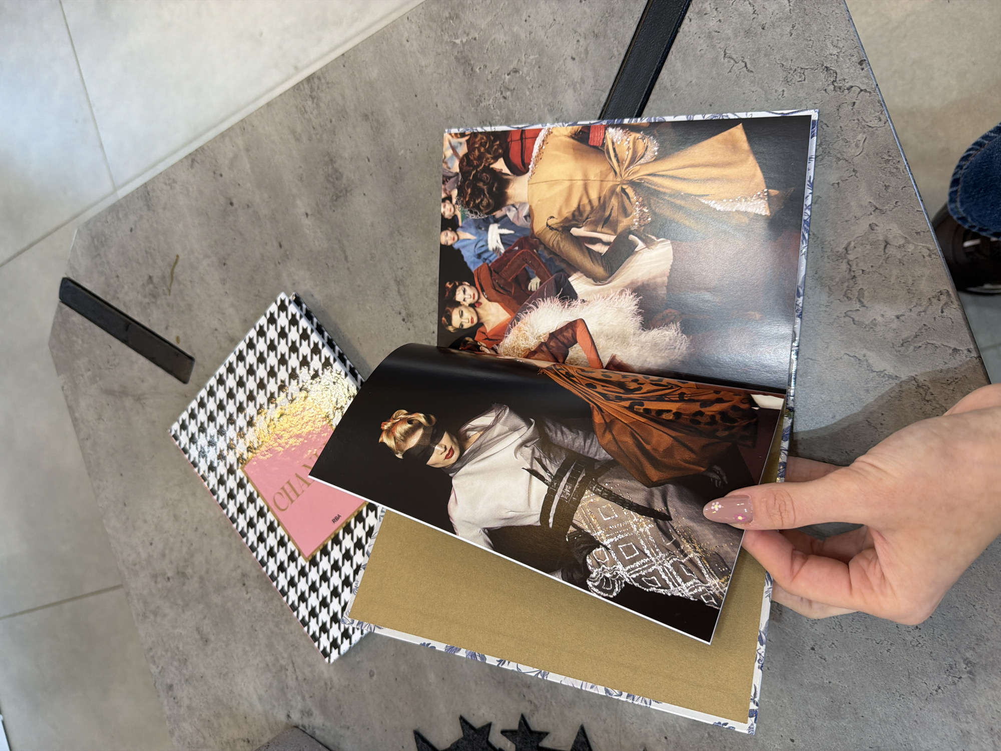 The image presents an overhead view of a table in an indoor setting, likely in Sofia, Bulgaria, showcasing fashion-related materials. A person's hand, with manicured nails featuring a delicate snowflake or floral design, is seen holding an open fashion magazine or photo book. The page held open displays a model wearing a black blindfold and an elaborate, textured white and silver garment, with a brown patterned fabric draped around them.
Adjacent to the open book, another closed book or box with a distinctive black and white houndstooth pattern is visible. A pink insert with gold accents and the partial text "CHAN" (likely "CHANEL") and "RBA" rests on top of this patterned item.
The table surface beneath the items is grey with a textured, stone-like or concrete finish. Parts of black table legs or supports are visible on the left and upper right. The background of the open book reveals another detailed fashion image featuring multiple models in ornate, historical or period-inspired clothing. A small portion of a person's leg in blue jeans and a dark shoe is visible on the far right. The scene depicts a casual moment of engaging with fashion media.