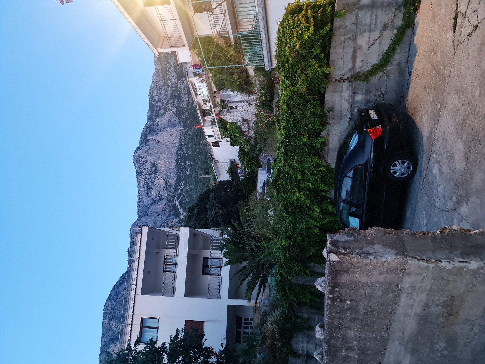 The image captures a view of a street in Brist, Croatia, at dusk. In the foreground, a concrete wall is visible, with a black sedan parked on a paved surface to its right. The car is facing away from the viewer, and its license plate is partially obscured. Beyond the parked car and the wall, a lush green hedge covers a concrete barrier.
Further up the slope, several white buildings with balconies are nestled against a backdrop of rugged, grey mountains. The sky above is a clear, pale blue, suggesting the late evening or early morning. A thin ray of light can be seen emanating from the upper right corner of the image. The overall scene depicts a residential area in a coastal town, characterized by its proximity to mountainous terrain.