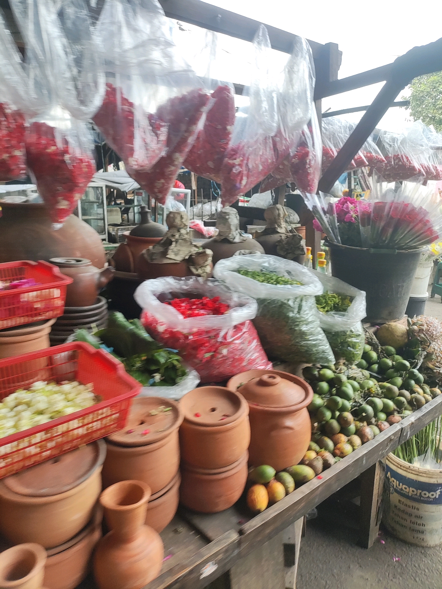 This image depicts an outdoor market stall in East Jakarta, Indonesia. The stall is filled with various goods, predominantly flowers and produce. Large plastic bags filled with red flower petals are hanging overhead. In the foreground, there are red plastic baskets containing what appears to be white chopped flowers and chopped green vegetables. Stacked terracotta pots of various sizes are also a prominent feature. To the right, a collection of green fruits, possibly betel nuts, are arranged on the stall, alongside some yellow and orange fruits. Behind these, a dark bucket holds several bouquets of pink and red flowers. Further back, more terracotta pots and what looks like a display of packaged goods can be seen. The scene is set against a backdrop of a wooden structure supporting the overhead coverings and some distant trees. The overall impression is that of a vibrant, bustling market.
