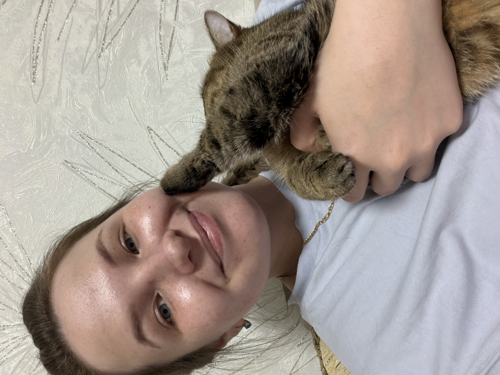 A woman lies on a surface decorated with a pale, textured pattern, likely a bed. A tabby cat lies on her chest, its paw touching her face. The woman is fair-skinned with light-colored eyes, and she's wearing a light-colored shirt. A gold necklace is visible. The location is Chaikovsky, Russia.
