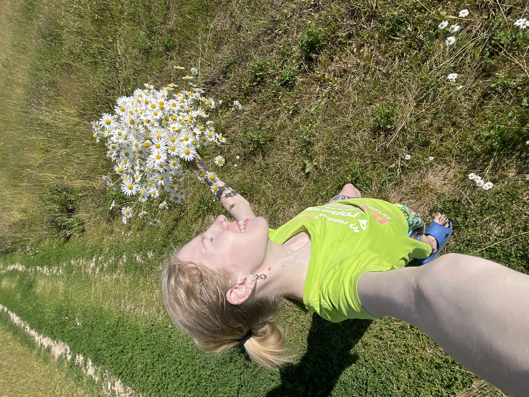 A young person with blonde hair, wearing a bright green t-shirt and blue sandals, lies on their back in a grassy field, holding a large bouquet of daisies. The person is looking upwards with their mouth open in what appears to be laughter or a shout of joy. Their hair is pulled back into a ponytail, and they have several piercings in their ears and a tattoo on their arm. The bouquet of daisies is voluminous and takes up a significant portion of the frame.
The setting is a sunny outdoor environment with a mix of green grass and drier, brown vegetation. Wild daisies are scattered throughout the field. In the background, a pathway or mowed strip of grass runs diagonally through the scene, suggesting a park or a rural area. The lighting indicates it is daytime, likely a sunny afternoon, given the shadows. The overall mood is cheerful and carefree, with the person enjoying the flowers and the natural surroundings. The text on the t-shirt is partially visible and appears to be in Cyrillic script. The location context is Сям-Можга, Russia.