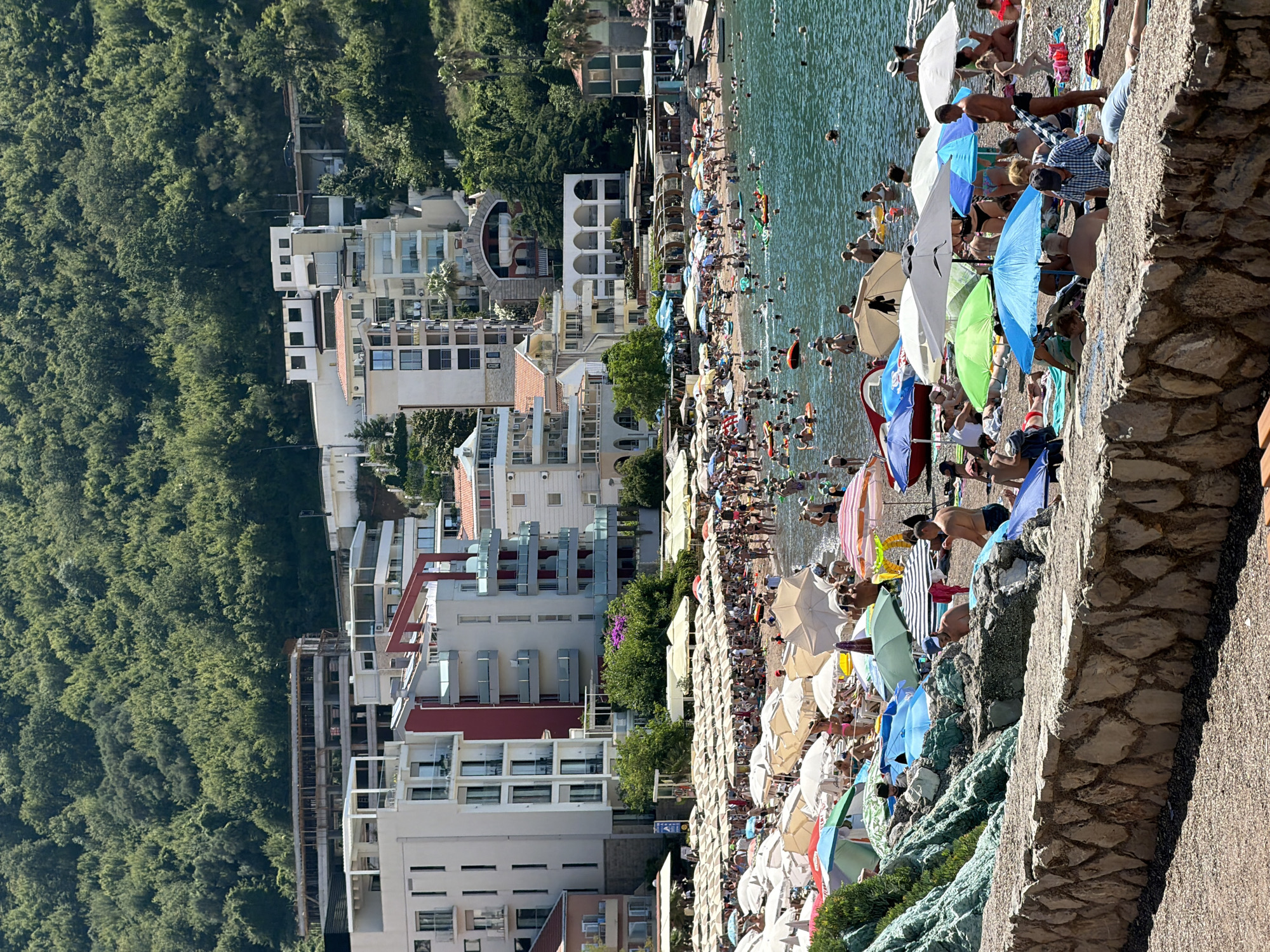 This is a medium-angle, outdoor, daytime shot of a crowded beach with a mountain slope covered in trees in the background. The beach is densely populated with people sunbathing and swimming. Numerous beach umbrellas, mostly white with some colorful ones, are spread across the sand, providing shade. The water is calm and blue, with many people in it, some on inflatable toys. Behind the beach, several multi-story buildings with white and red facades are visible, along with some construction. A stone wall forms a barrier in the foreground. The overall scene depicts a bustling summer day at a beach resort.
