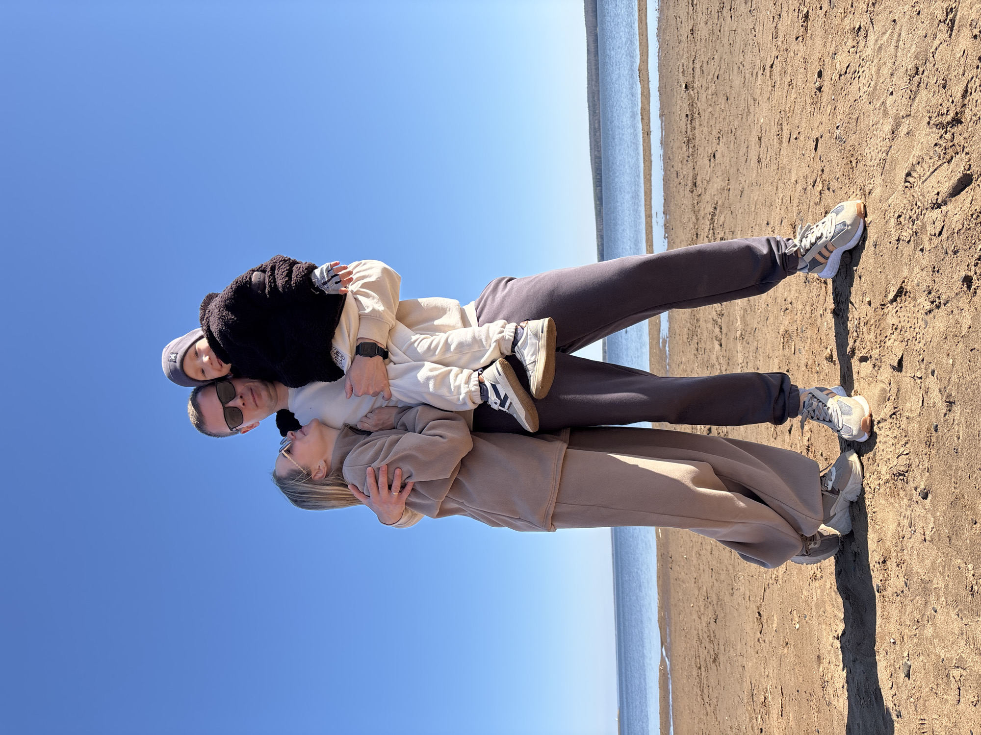 A family of three stands close together on a beach in Chaikovsky, Russia, under a clear blue sky. The photo appears to be taken from a low angle. The beach has light brown sand, bordering a body of water, likely a river or lake. The family consists of a woman, a man, and a small child. The woman wears a light brown tracksuit and sunglasses. She is hugging the man. The man also has sunglasses and dark-colored pants. He is holding the child, who is dressed in light-colored clothing and a dark jacket. The family's feet are partially visible, showing their footwear on the sand.