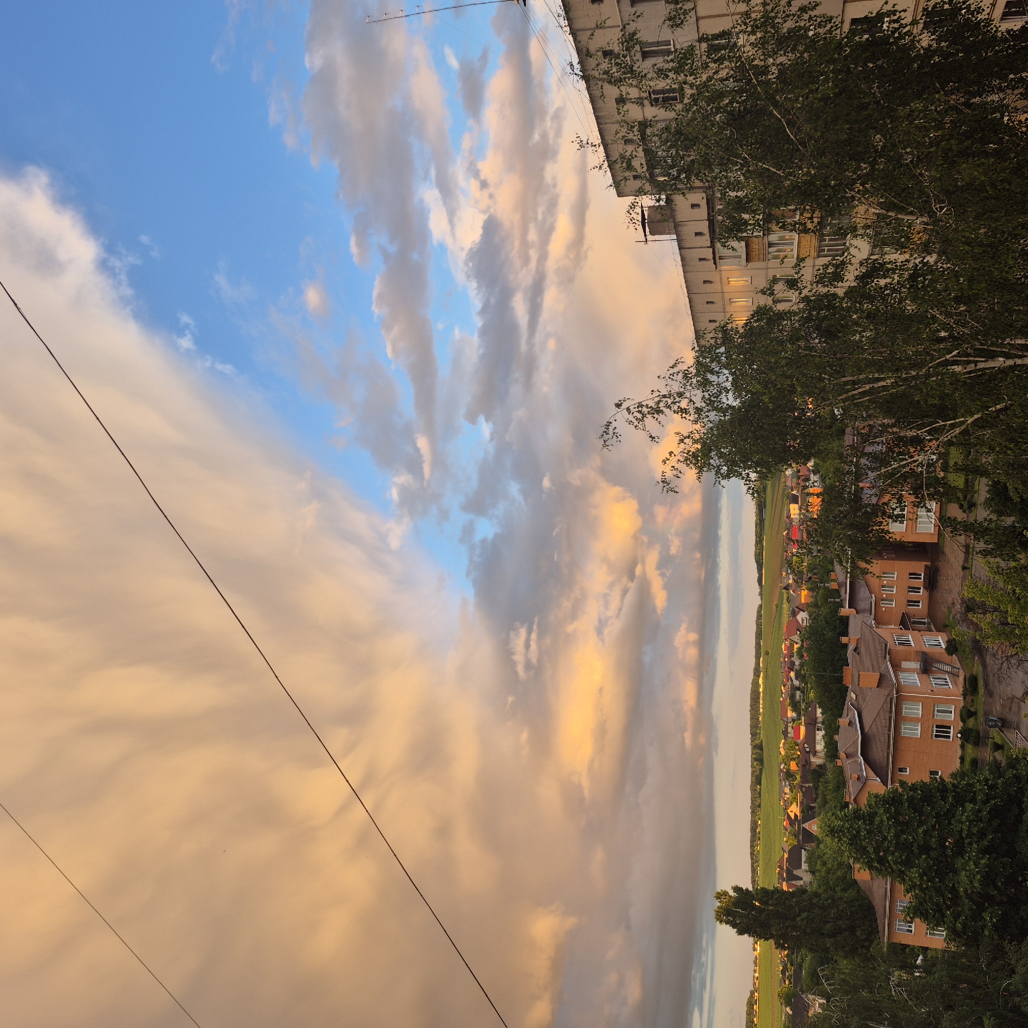 The image captures a panoramic view of a residential area in Bila Tserkva, Ukraine, at dusk. A dramatic sky dominates the upper portion of the frame, with large, voluminous clouds tinged with warm hues of orange and yellow from the setting sun, contrasting with patches of clear blue sky. Two thin, black electrical wires stretch diagonally across the sky, one from the upper left to the middle right, and the other from the upper left to the lower left.
In the foreground and midground, a mix of buildings and greenery is visible. On the right, a multi-story apartment building with a neutral-colored facade and visible windows stands prominently. In front of this building, and extending towards the center of the image, is a cluster of deciduous trees, their leaves a vibrant green. To the left of the apartment building, and further into the midground, are several smaller residential buildings with terracotta-tiled roofs. These appear to be houses, each with multiple windows and some with balconies. A tall, slender evergreen tree stands out amongst the houses on the left side of the image.
The background reveals a sprawling landscape with more houses, their roofs a mix of colors, nestled among trees. Beyond the residential areas, rolling green fields stretch towards the horizon, bathed in the soft light of the setting sun. The overall impression is one of a calm evening scene in a suburban or semi-urban environment. No people or animals are discernible in the image. The ground appears to be damp, possibly from recent rain, particularly noticeable in the paved areas between the houses on the left.