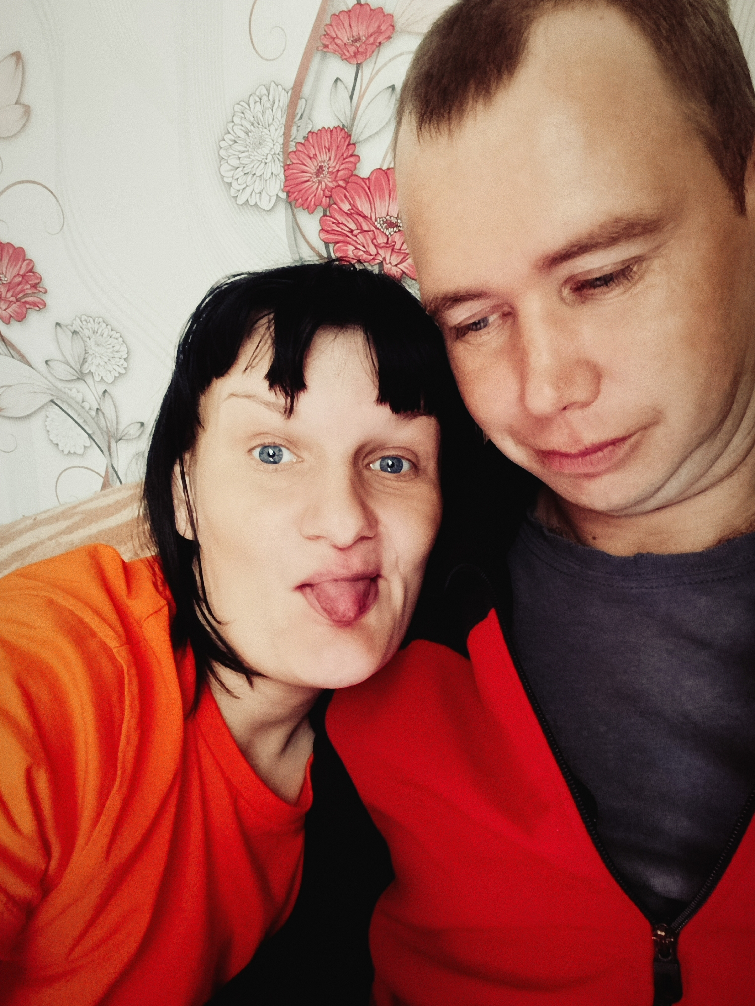 A close-up shot depicts a man and a woman posing for a selfie against a background of floral wallpaper. The woman on the left wears an orange shirt and has short, dark hair with bangs. Her blue eyes are wide open, and she sticks out her tongue playfully. The man on the right has short, light brown hair and wears a gray shirt with a red jacket partially visible. He looks down with a slight frown. The location is unknown, in the city of Unknown, Unknown.