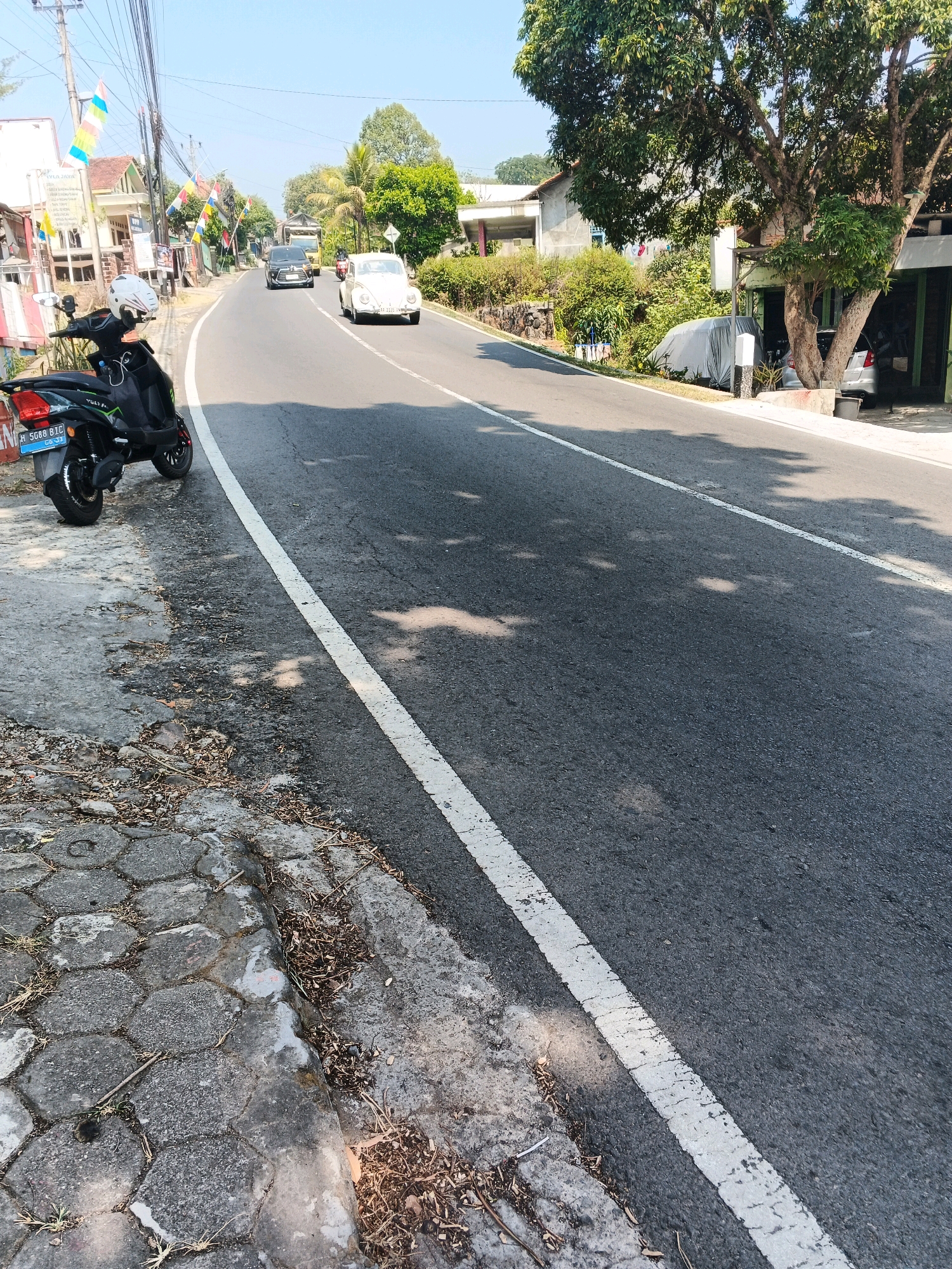 The image depicts a street scene in Pakopen, Indonesia. A dark-colored scooter with a white helmet resting on its seat is parked on the left side of the road. The road itself is paved and marked with a white line. Several vehicles, including a white classic car and a black SUV, are traveling on the road. The surroundings include residential buildings, trees, and vegetation. The sky is clear and blue, indicating a sunny day. The pavement on the side of the road is made of interlocking hexagonal concrete blocks.