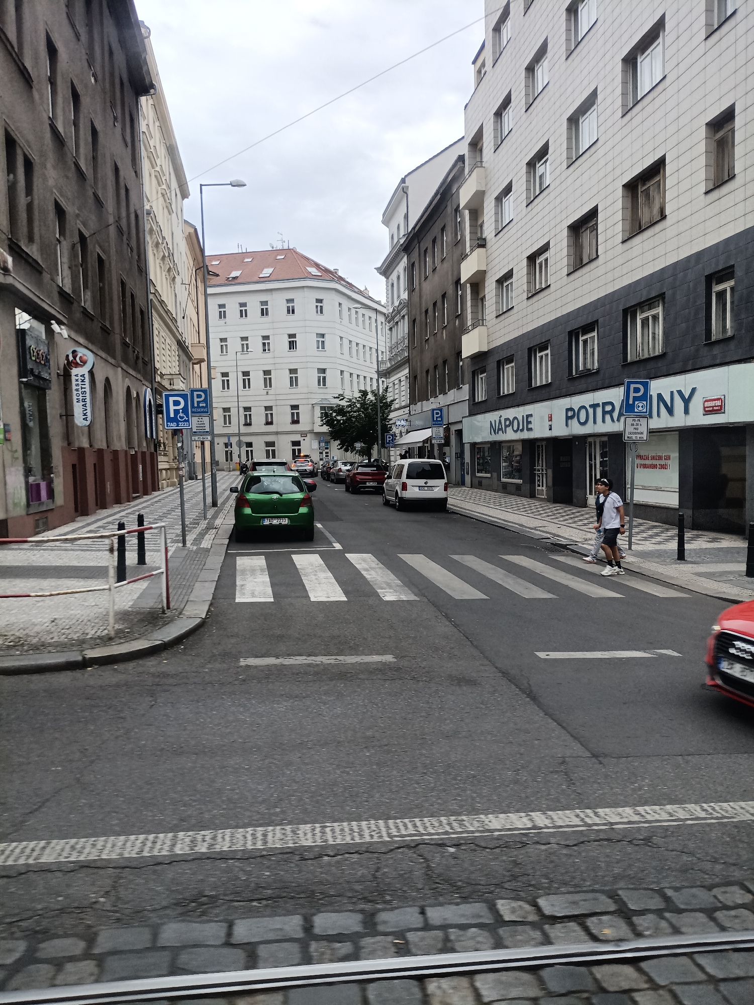 This is a street-level, medium-range, eye-level shot taken in Prague, Czechia, on an overcast day. The scene depicts a narrow city street with buildings on either side. The foreground shows tram tracks embedded in cobblestone pavement. A pedestrian crossing with white stripes is visible on the asphalt roadway.
On the left side of the street, a multi-story building with a dark facade and numerous windows dominates the view. A storefront with the text "MORSKA AKVARISTIKA" is present. Several parked cars are on this side of the street, and a green hatchback is stopped at the pedestrian crossing. Two blue parking signs are mounted on a pole, one with a wheelchair symbol.
On the right side of the street, a modern building with a grey tiled facade and dark lower section stands tall. It features numerous windows and balconies. Below the building, storefronts with signage such as "NÁPOJE POTRAVINY" are visible. Two individuals, a younger person in a white t-shirt and dark shorts, and another person behind them in a dark t-shirt and shorts, are crossing the street at the pedestrian crossing. A white minivan is parked further down the street, and a red car is partially visible in the lower right corner of the frame.
The street is lined with streetlights and bollards. The buildings suggest a commercial and residential area. The overall atmosphere is that of a typical European city street.