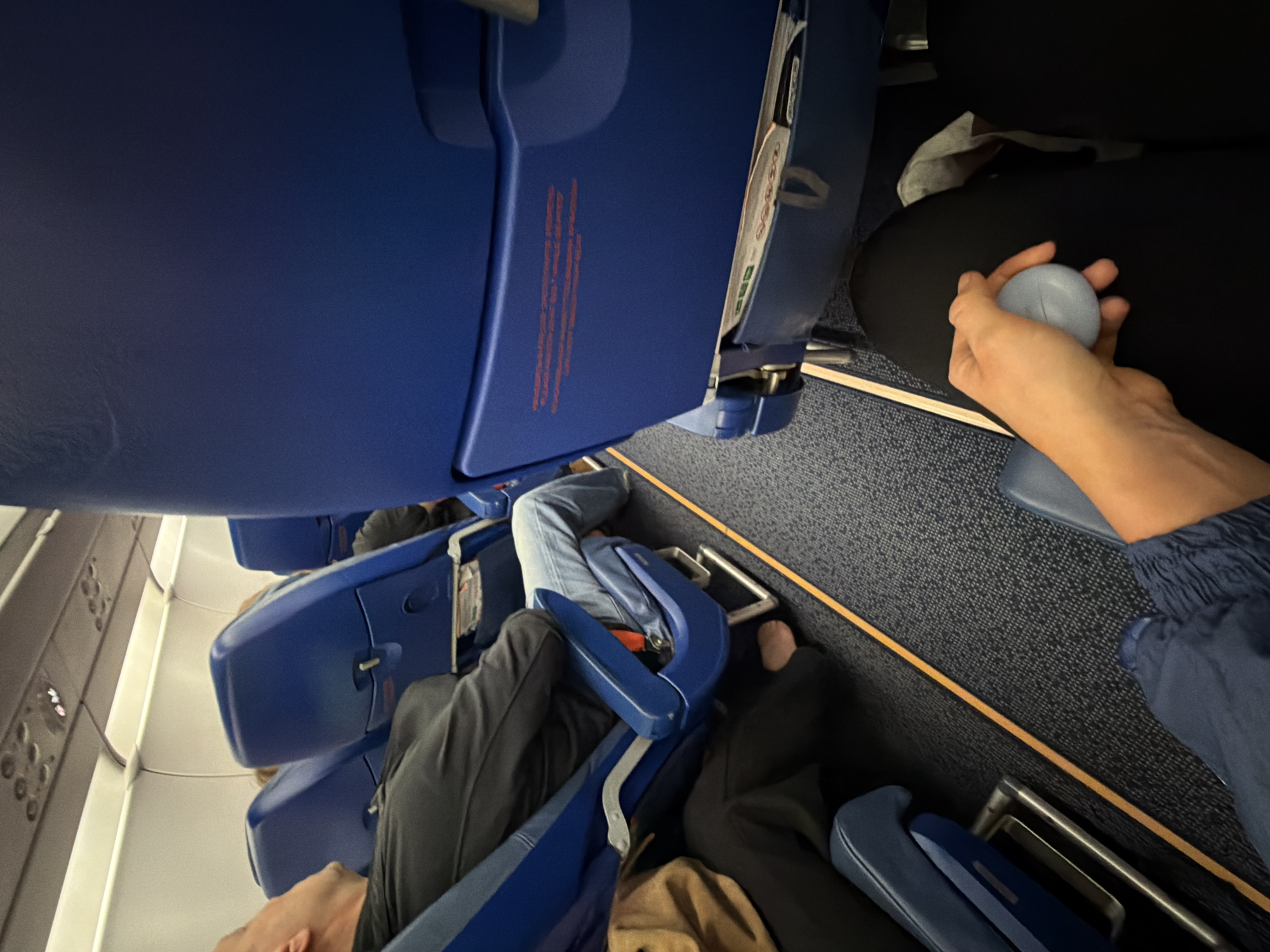 The image depicts the interior of an airplane cabin with blue seats. In the foreground, a person's hand is holding a small, light blue object. To the left, a man is seated with his eyes closed, appearing to be asleep. Another passenger is seated to his left, partially visible. The cabin floor is carpeted in a dark gray pattern with an orange line running along the aisle. The overhead compartment and lighting are visible at the top of the image. The scene is an everyday occurrence on commercial air travel. This image does not contain any information that would indicate a specific location in Saint Petersburg, Russia.