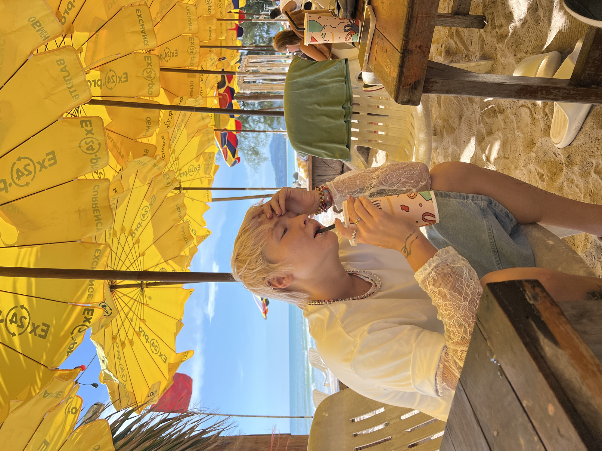 A young person with short blonde hair is sitting on a beach chair under a yellow umbrella, drinking from a pink cup with a straw. They are wearing a white t-shirt with white lace sleeves, denim shorts, and a beaded necklace. The person has a tattoo on their arm and is wearing rings on their fingers. The background shows a sandy beach with more yellow umbrellas, chairs, a table, and the ocean. The sky is blue with some clouds.