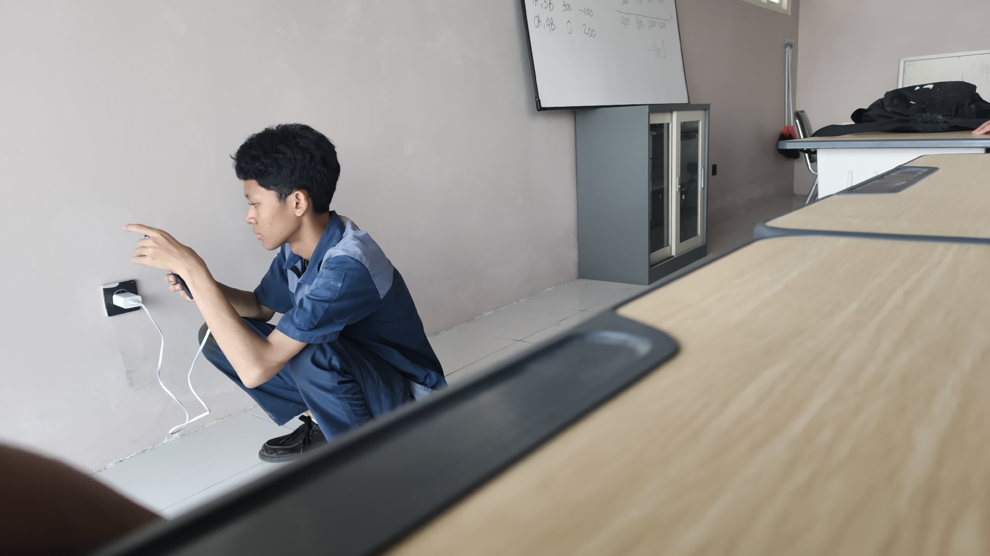 A young man in a blue work uniform is crouching next to a wall in what appears to be a classroom or office setting. He is plugging a white charger into a wall socket. The wall is a pale pink color, and the floor is tiled with white squares. In the background, there is a whiteboard with writing on it, a grey cabinet, and several tables. The tables have light wood surfaces and dark grey edges. One table is in the foreground, blurring the image. The lighting suggests it is daytime, with natural light coming from unseen windows. The man's posture and focused expression indicate he is engaged in the task of connecting the charger. The overall scene is one of everyday activity in a functional indoor space. The location is identified as Katrungan, Indonesia.