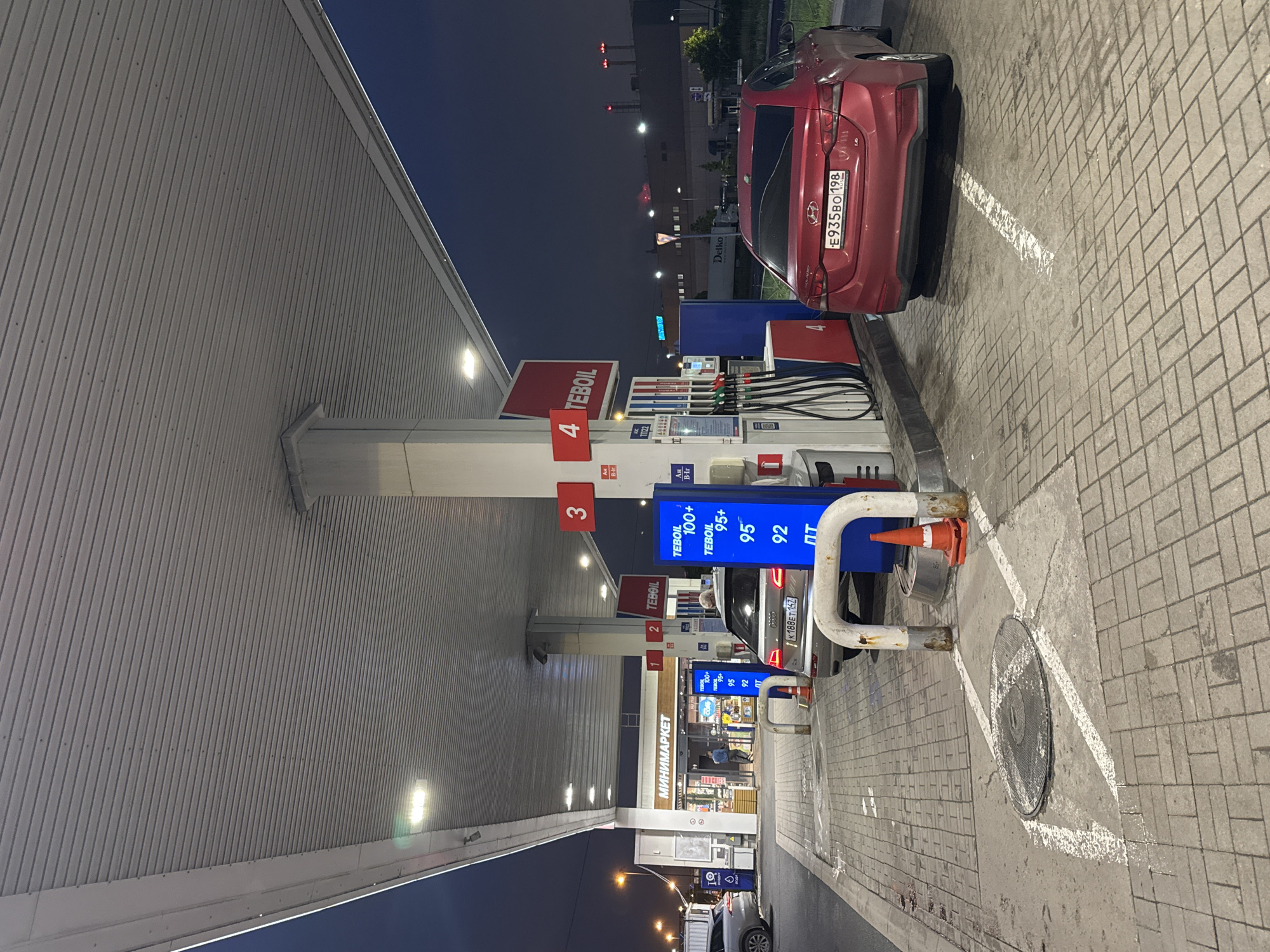 The image captures a TeBoil gas station in Saint Petersburg, Russia, at dusk or night. The station features multiple pumps under a white, corrugated metal awning. Prices for different grades of fuel are displayed in blue digital signs: TeBoil 100+, TeBoil 95+, 95, and 92. A minimarket building is visible behind the pumps. A red car is parked at the pump numbered '4' with a license plate E935BO198. Another car, a silver sedan with the license plate K188ET 147, is near the pump numbered '2'. The ground is paved with grey bricks. Orange traffic cones are positioned around a metal pipe near a manhole cover painted with white lines. Other details, such as the Delko company signage in the background, suggest a commercial or industrial area.