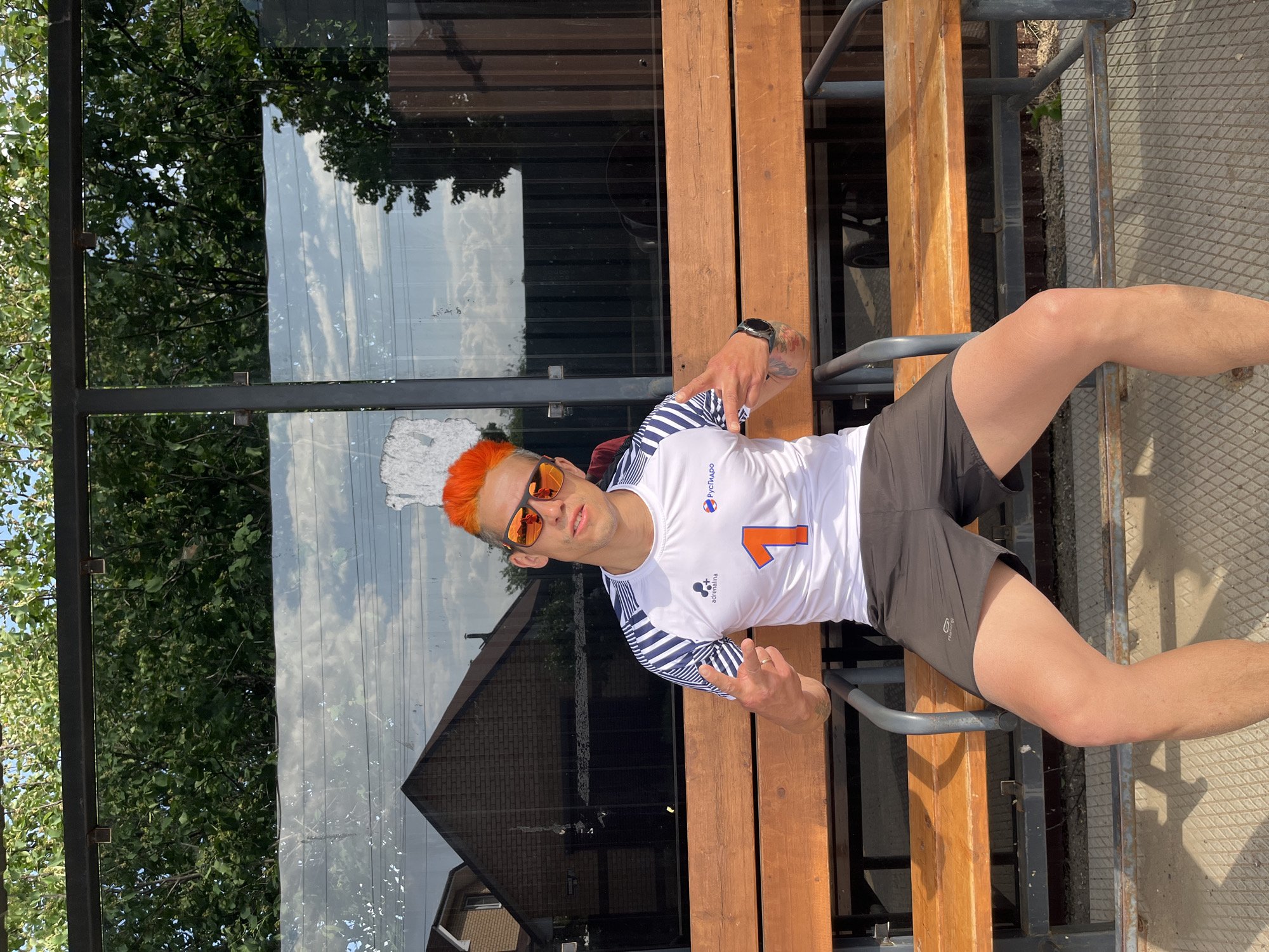 A man with orange hair and sunglasses is sitting on a wooden bench in Chaikovsky, Russia. He's wearing a white athletic shirt with blue stripes and the number "1" printed on it, along with grey shorts. He is making hand gestures with both hands, seemingly pointing. The bench is part of a structure with metal supports, and behind him is a glass panel reflecting trees and buildings. The scene appears to be outdoors during the daytime.