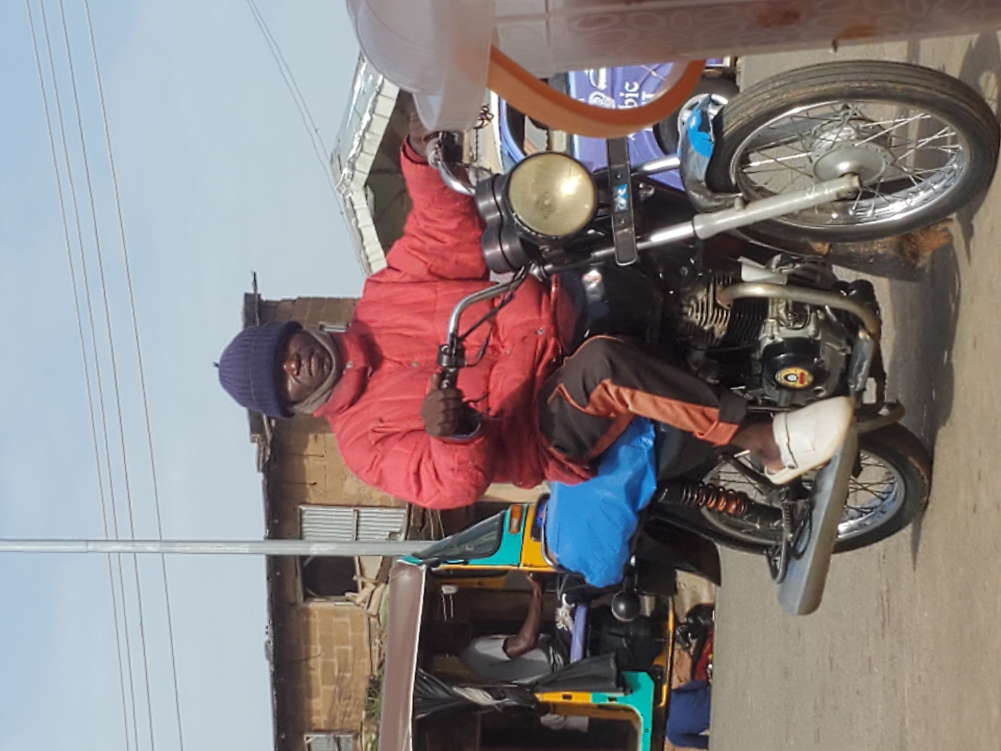 The image shows a man on a motorcycle in Gwong, Nigeria. The man is wearing a red jacket, a blue beanie, and white sneakers. He appears to be looking at the camera. The motorcycle is black with chrome accents. In the background, there is a building with a corrugated metal roof and what appears to be a tuk-tuk. The sky is blue and there are power lines visible. The time of day appears to be daytime, and the weather seems to be clear. The scene is a street in a populated area, likely a market or a residential street. The man's expression is neutral, and there is no clear indication of an emotion. There is no visible text in the image.