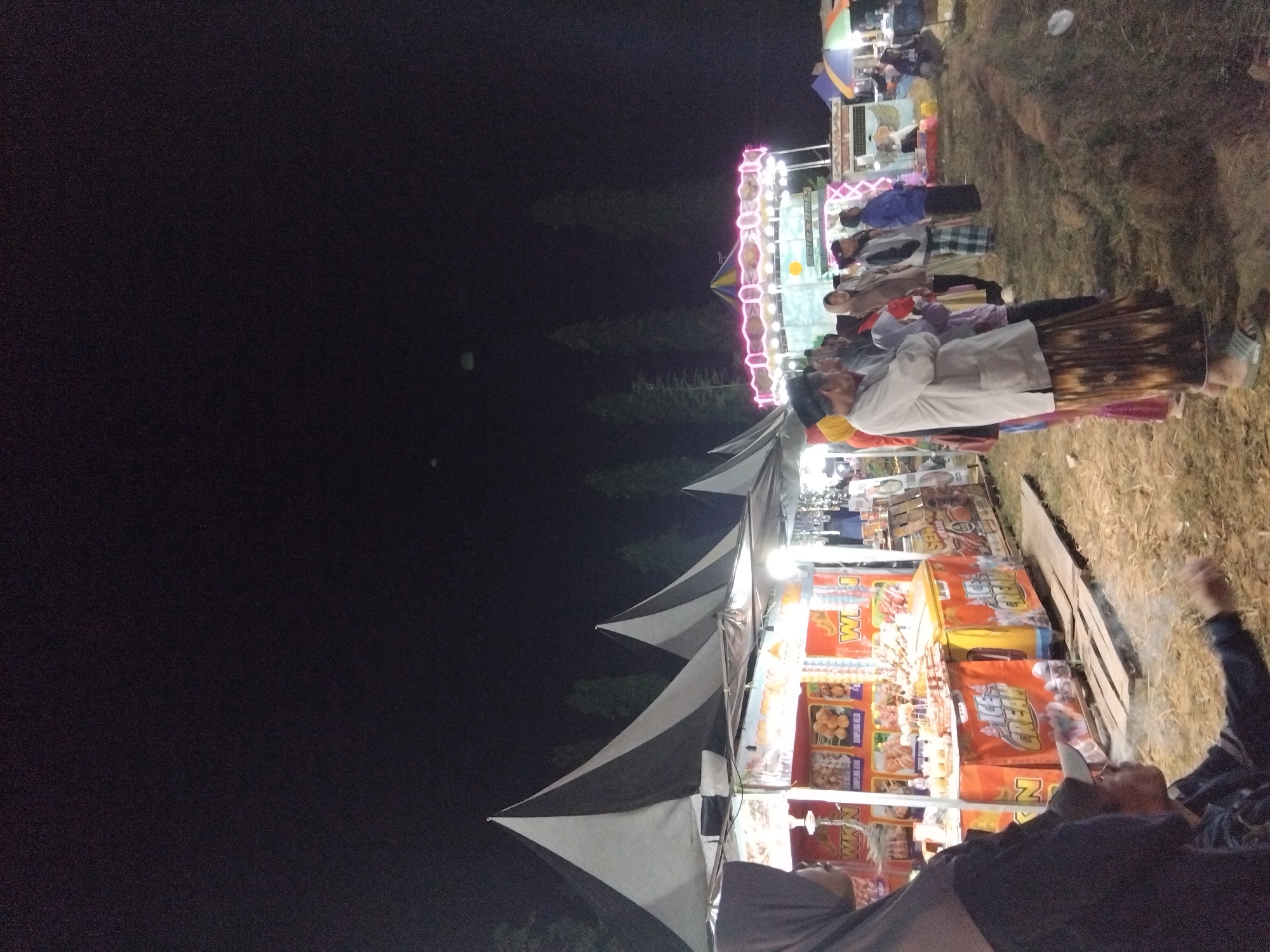 An outdoor nighttime market scene is depicted in Sambirata, Indonesia. In the foreground, a person wearing a black jacket and a grey baseball cap is sitting and smoking, with smoke rising around them. To the left, a person wearing a dark headscarf is partially visible. Behind them, a food stall displays various items including "Ice Cream" signage, and pictures of food items like dumplings. Further back, a group of people are standing and observing. A carousel with colorful lights is visible in the background, illuminated against the dark night sky. The ground is covered in dry grass and dirt.