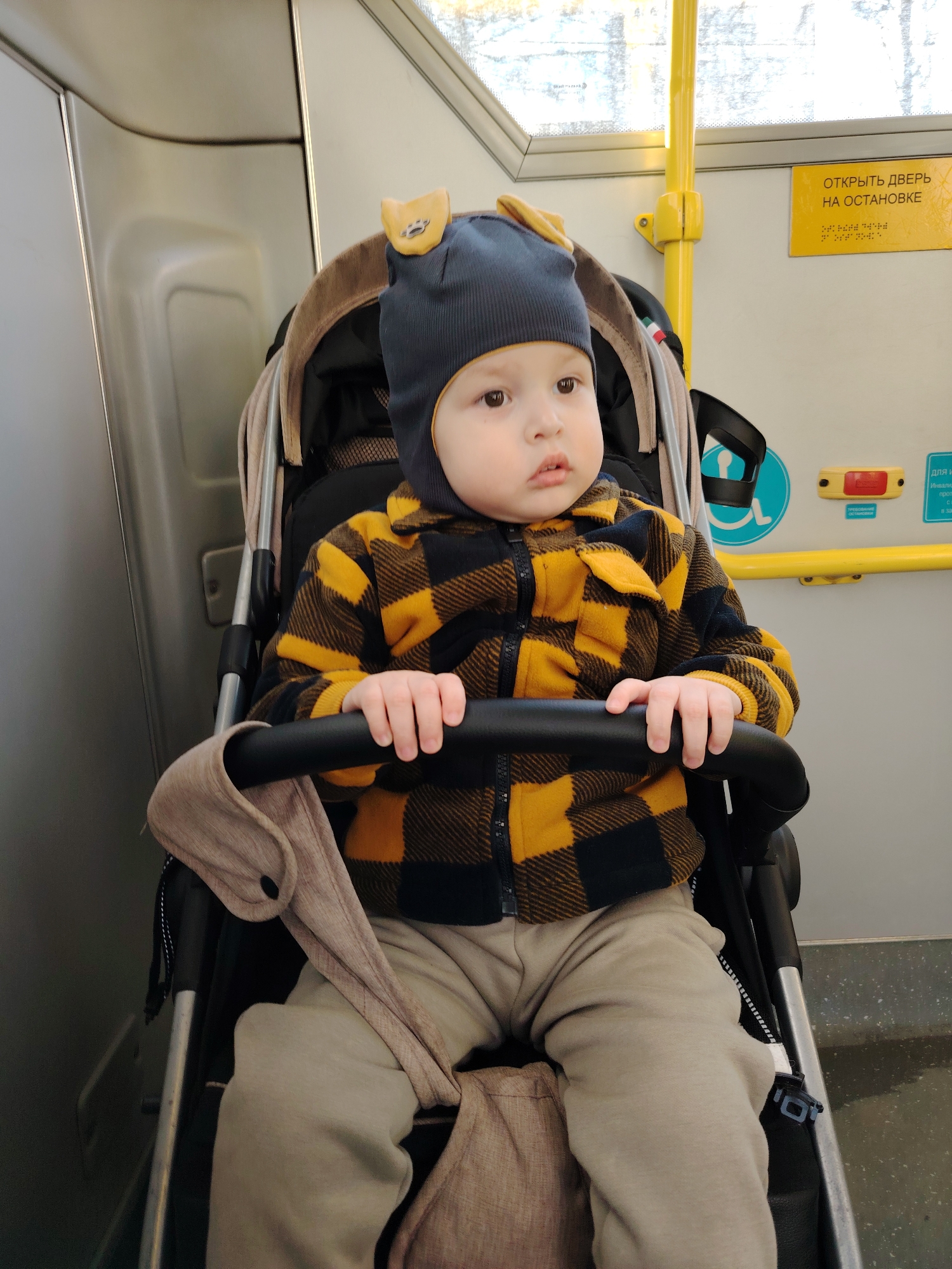 A young child is seated in a beige and black stroller inside a public transport vehicle, likely a bus or tram, in Chaikovsky, Russia. The child, an infant or toddler, wears a dark blue ribbed beanie with two tan, ear-like protrusions featuring a dark paw print, a black and yellow plaid zip-up jacket, and light tan sweatpants. Their hands rest on the stroller's black bumper bar as they look slightly to the right with a neutral expression. The vehicle's interior features light gray/beige wall and ceiling panels, a bright yellow vertical handrail on the right, and a window revealing a blurred outdoor scene. A yellow rectangular sign on the upper right wall displays black Cyrillic text, "ОТКРЫТЬ ДВЕРЬ НА ОСТАНОВКЕ" (OPEN THE DOOR AT THE STOP), with Braille below it. A light blue circular accessibility symbol with a white wheelchair icon and a red stop request button are partially visible on the wall to the right of the child.