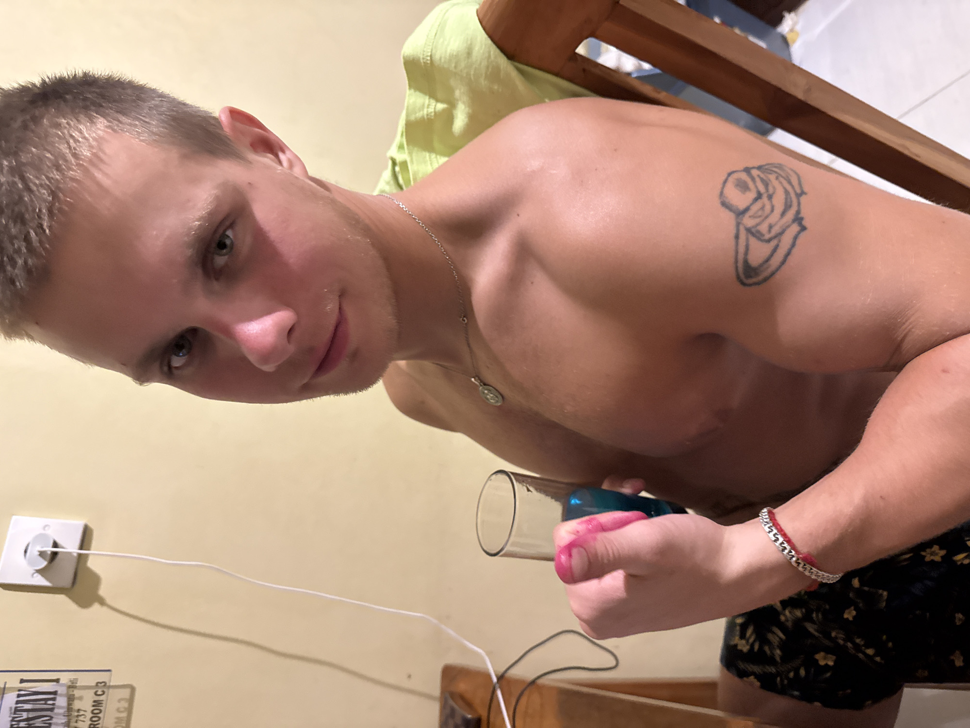 A young adult male is positioned indoors, facing the camera, with the perspective being a low-angle shot. He is shirtless, exposing a tattoo on his upper left arm, and wears a silver chain necklace. He is holding a clear glass test tube in his left hand with a blue syringe inserted, filled with pink liquid. The background includes a light green cloth draped over what appears to be a wooden chair, a white wall with a wall outlet, and part of a decorative sign. He is wearing black boxer shorts with floral print. This scene is likely located in a room in Kuta Selatan, Indonesia.