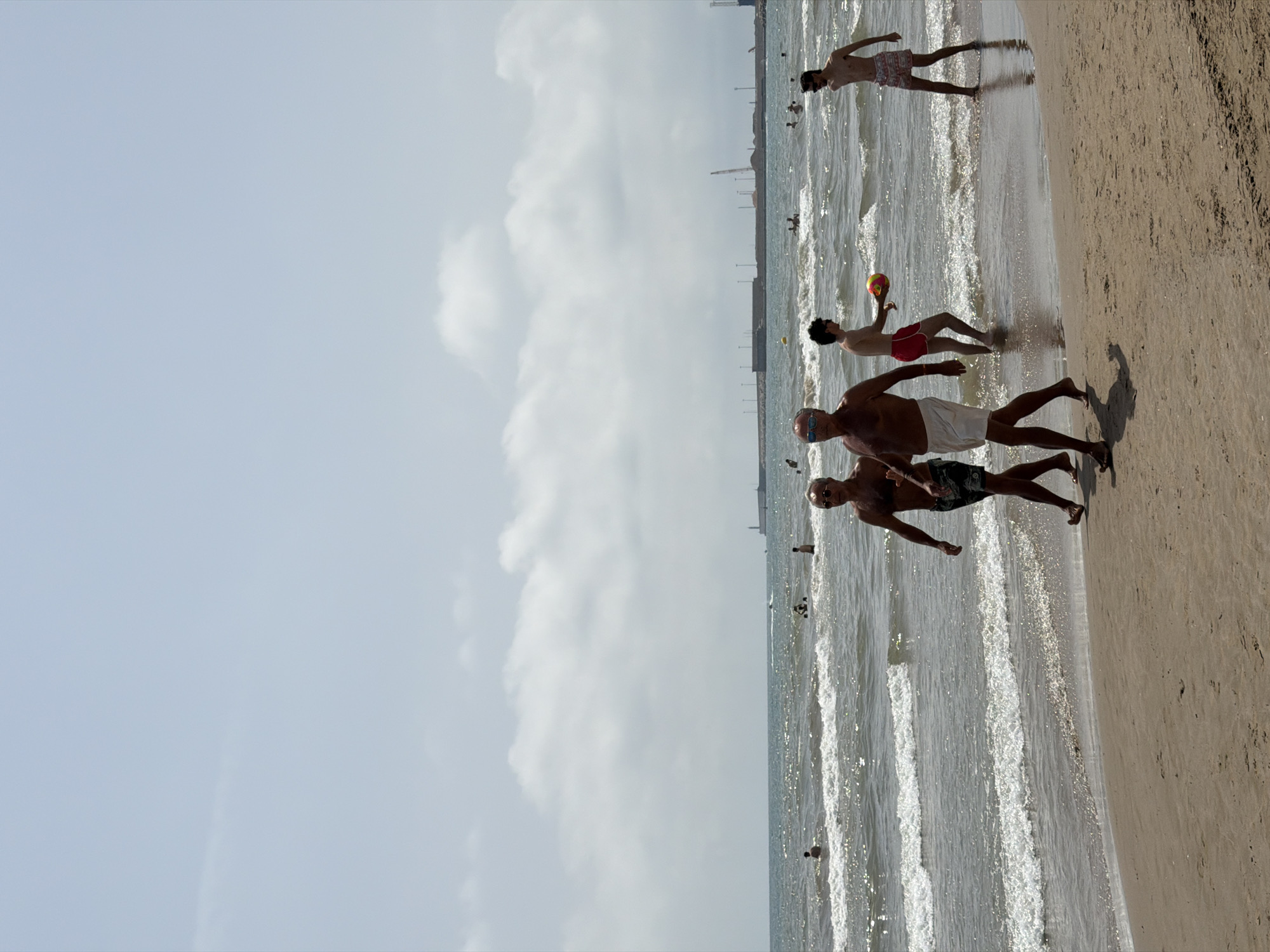 The image depicts a sandy beach on a cloudy day. The ocean is visible in the background with waves and several people swimming or wading in the water. In the foreground, four men are walking on the beach. Two of the men are walking together, wearing swim trunks. Another man is walking slightly behind them, also in swim trunks, and is holding a colorful ball. The fourth man is walking towards the right of the frame, wearing patterned swim trunks. In the far background, there is a pier or breakwater with structures and cranes, suggesting a port or industrial area. The sky is overcast with some clouds.