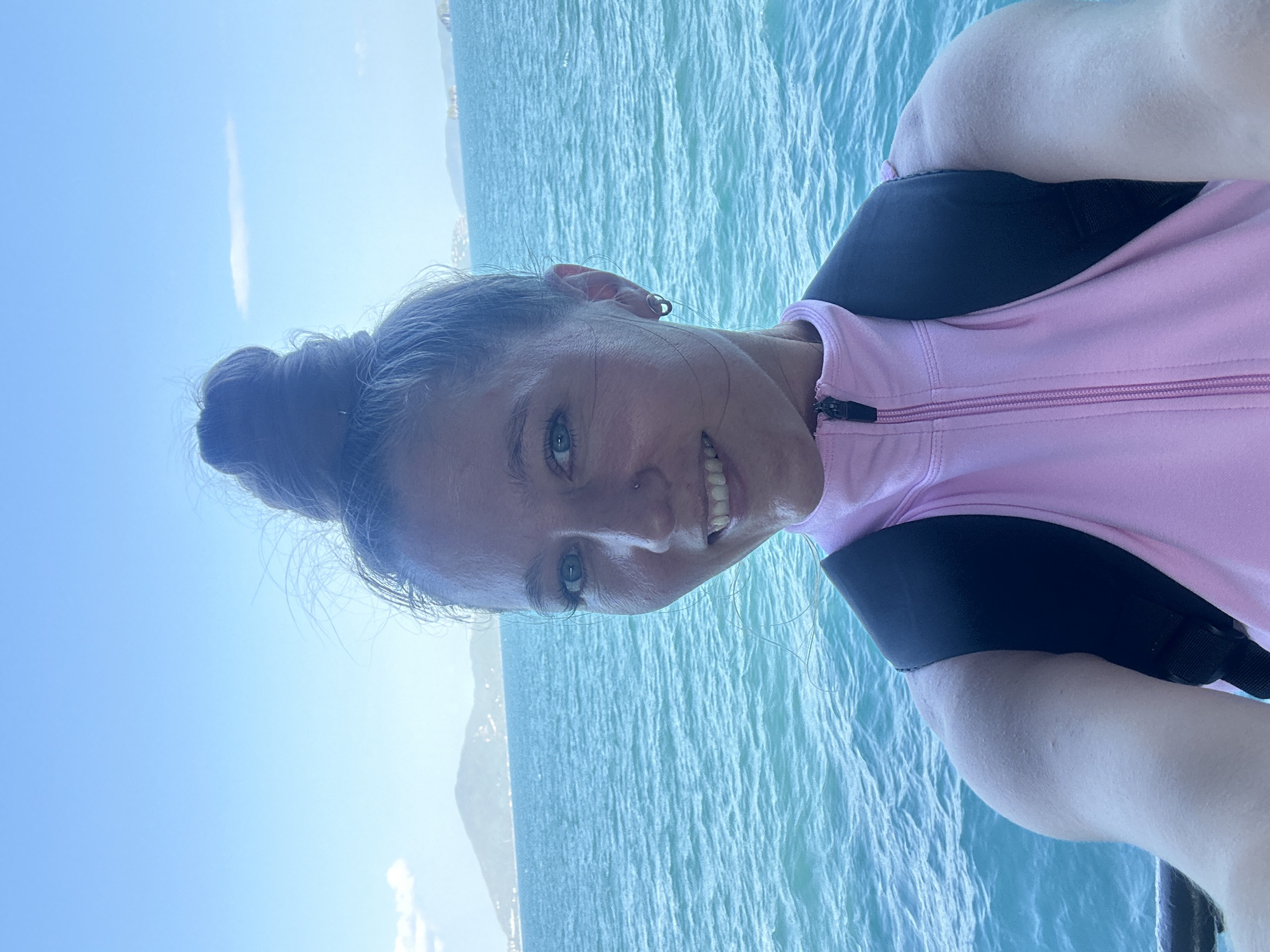 A young woman with blonde hair pulled up in a bun and blue eyes is taking a selfie with a backpack on. She is wearing a pink zip-up shirt. She is smiling at the camera. The background is the ocean with islands in the distance and a blue sky. The location is likely a ferry or boat in Thailand.