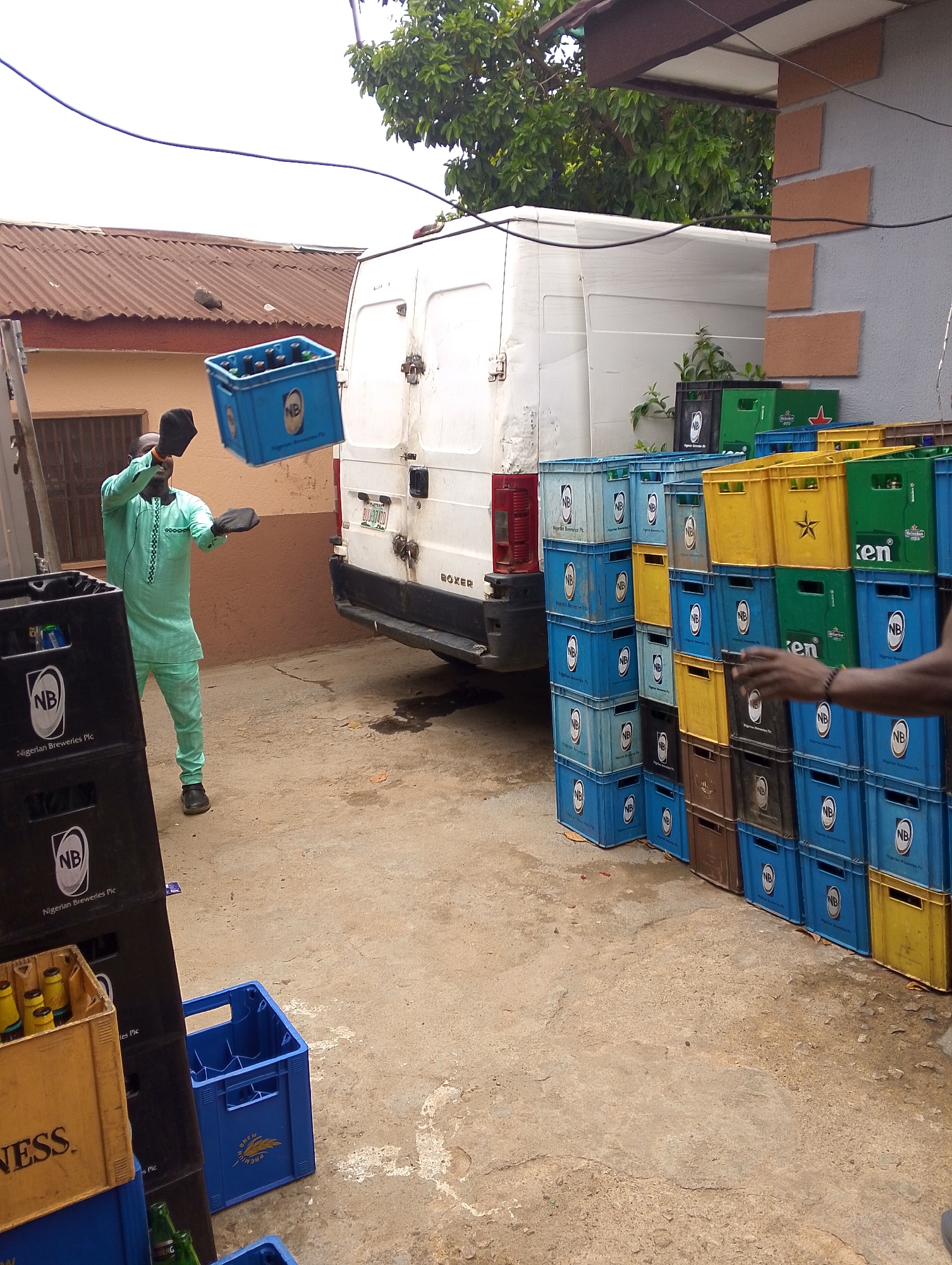 This image captures an outdoor scene of beverage handling, likely at a distribution point or shop in Jos, Nigeria. In the foreground, a man dressed in a light green traditional tunic and black cap is engaged in active labor. He is caught mid-motion, seemingly tossing or catching a blue plastic crate filled with glass bottles, which is suspended in the air. The crate prominently displays the "NB Nigerian Breweries Plc" logo. Another person's arm and hand are visible on the right, suggesting involvement in the same task of moving crates.
The setting is an unpaved yard, likely a service area, with a white delivery van, a "BOXER" model with a Nigerian license plate "PLT 574TD," parked in the background. Abundant beverage crates are stacked on the right side of the frame. These include numerous blue crates marked "NB Nigerian Breweries Plc," black crates also bearing the "NB Nigerian Breweries Plc" and "Guinness" logos, yellow crates (some with a star symbol, likely for Star Lager), and green crates branded "Heineken" (with "Ken" clearly visible). Some crates contain bottles, while others are empty.
Notable details include a building with a corrugated iron roof on the left and a wall with decorative orange rectangular accents on the right. Overhead, power cables stretch across the frame. The daylight appears bright but diffused by an overcast sky. The visible text on the crates and the van's license plate, along with the man's attire, strongly indicate a Nigerian context, aligning with the provided location of Jos. The scene depicts a routine logistical activity involving the movement and organization of bottled beverages.