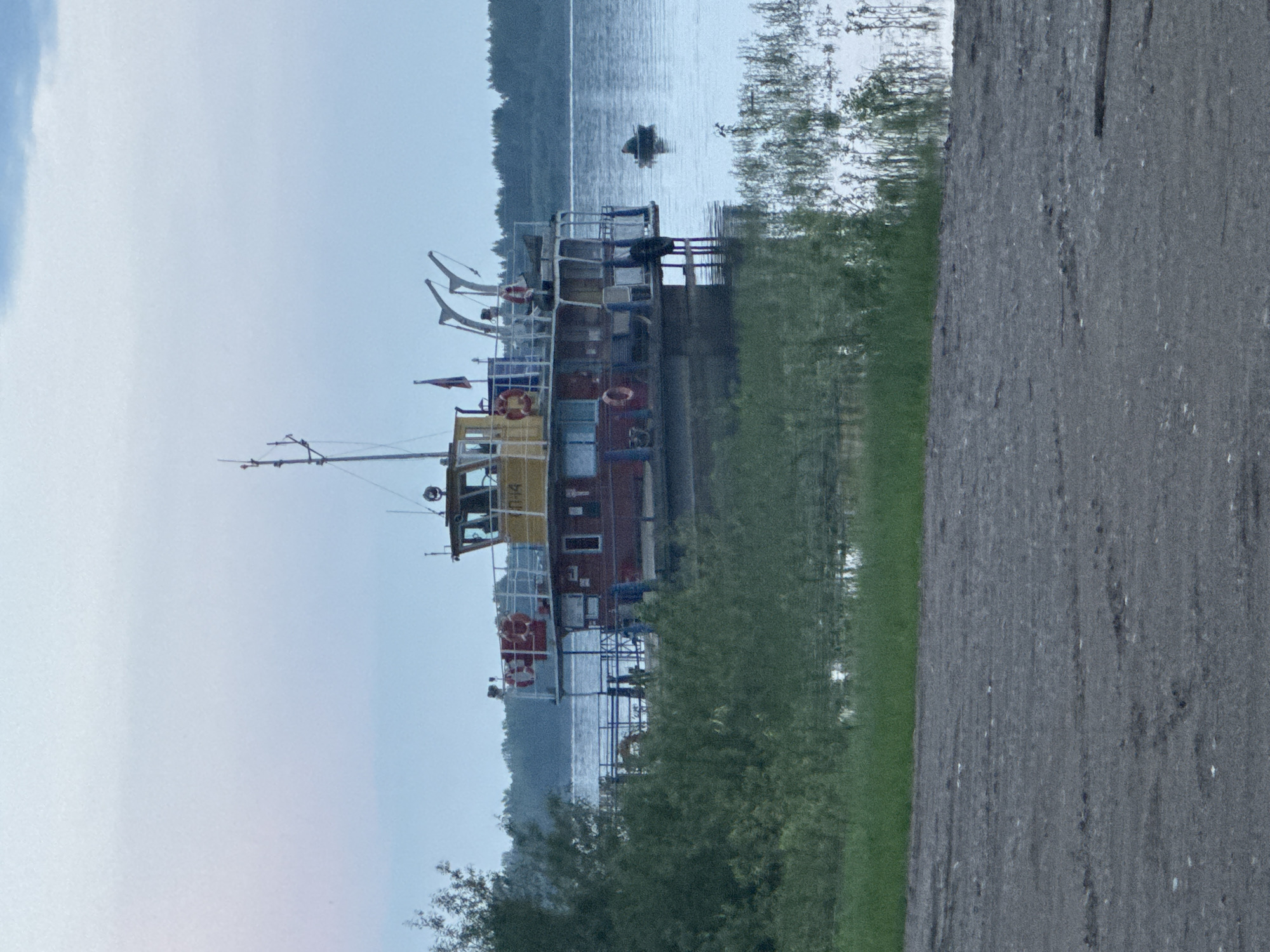 An overcast, hazy daytime scene depicts a river with a tugboat moored at a dock. The tugboat is red with a yellow cabin, and it has a life preserver and two flags on its deck. In the distance, a lone figure in a small boat is visible on the water. The foreground shows a dirt path and green vegetation. The location is in the city of Tyulkino, Russia.