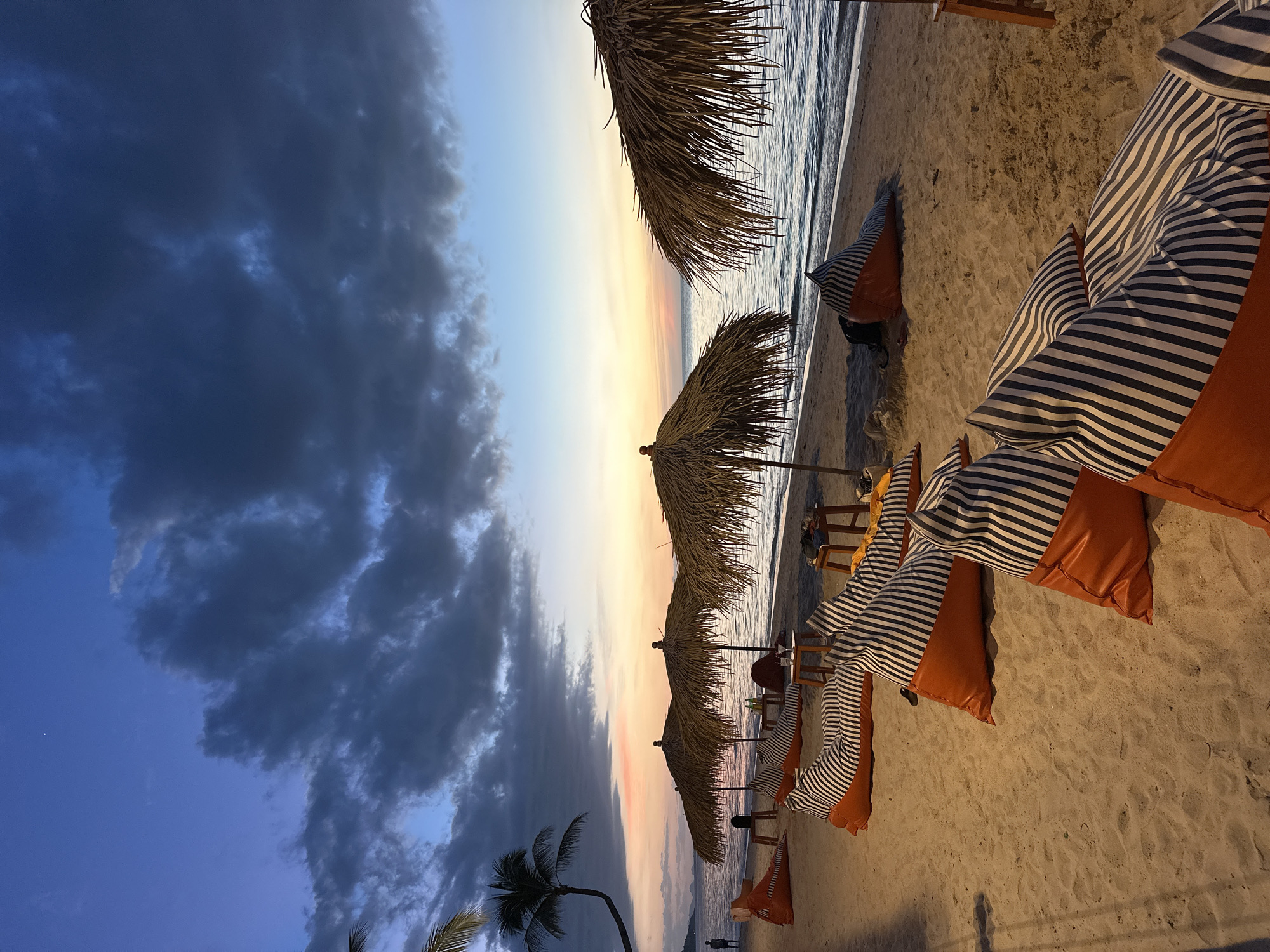 A serene beach scene unfolds at dusk in Ko Samui District, Thailand, with a twilight sky painted in hues of deep blue, violet, and soft orange. The calm ocean gently laps the sandy shore, reflecting the fading light.
Arranged on the sand are several large, orange beanbag chairs adorned with black and white stripes, some unoccupied and others appearing to be in use. In front of them are low wooden tables, some holding drinks and a cocktail glass.
Several thatched umbrellas provide shade, their rustic texture contrasting with the smooth sand. In the distance, a lone figure stands near the water's edge. A palm tree with a gracefully curved trunk leans over the beach, its fronds silhouetted against the colorful sky. The overall atmosphere is tranquil and inviting, suggesting a peaceful evening by the sea.