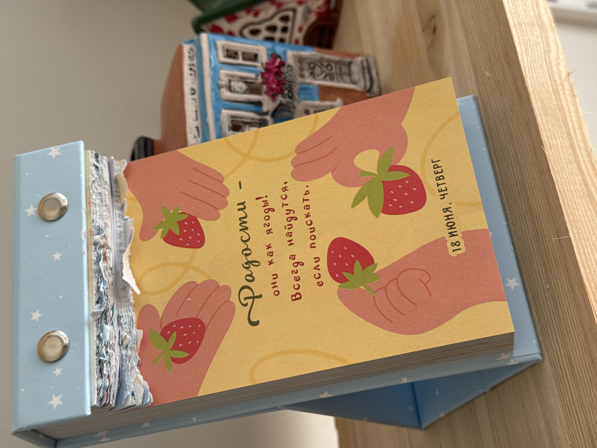 The image shows a close-up of a flip calendar standing on a wooden shelf. The calendar has a light blue cover decorated with white stars and two silver fasteners. The pages of the calendar are torn at the top, revealing layers of paper. The main page displayed has a yellow background and features an illustration of several hands holding strawberries. Text in Russian is printed on the page, and below the illustration, the date "18 ИЮНЯ, ЧЕТВЕРГ" (June 18, Thursday) is visible. In the background, out of focus, are miniature decorative houses, one of which is blue and orange with windows and flowers. The shelf appears to be made of light-colored wood. There is no discernible connection to the city of Izhevsk, Russia.
