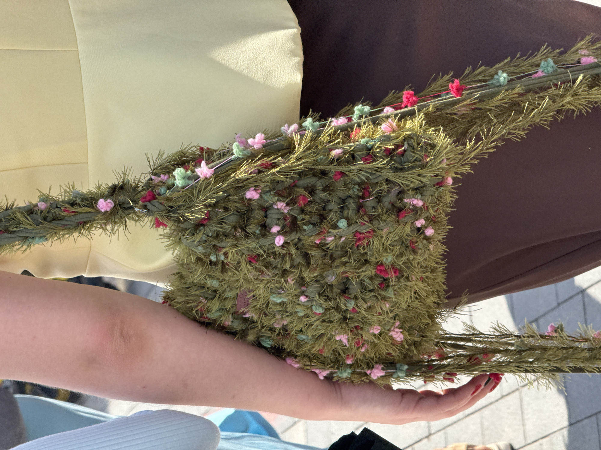The image shows a close-up of a person's arm holding a green, textured bag adorned with small pink and blue pom-poms and floral accents. The bag has a string strap. The person is wearing a light yellow top and brown pants. The background appears to be an outdoor setting with a paved surface and some greenery visible. There is no indication of the location being Yekaterinburg, Russia.