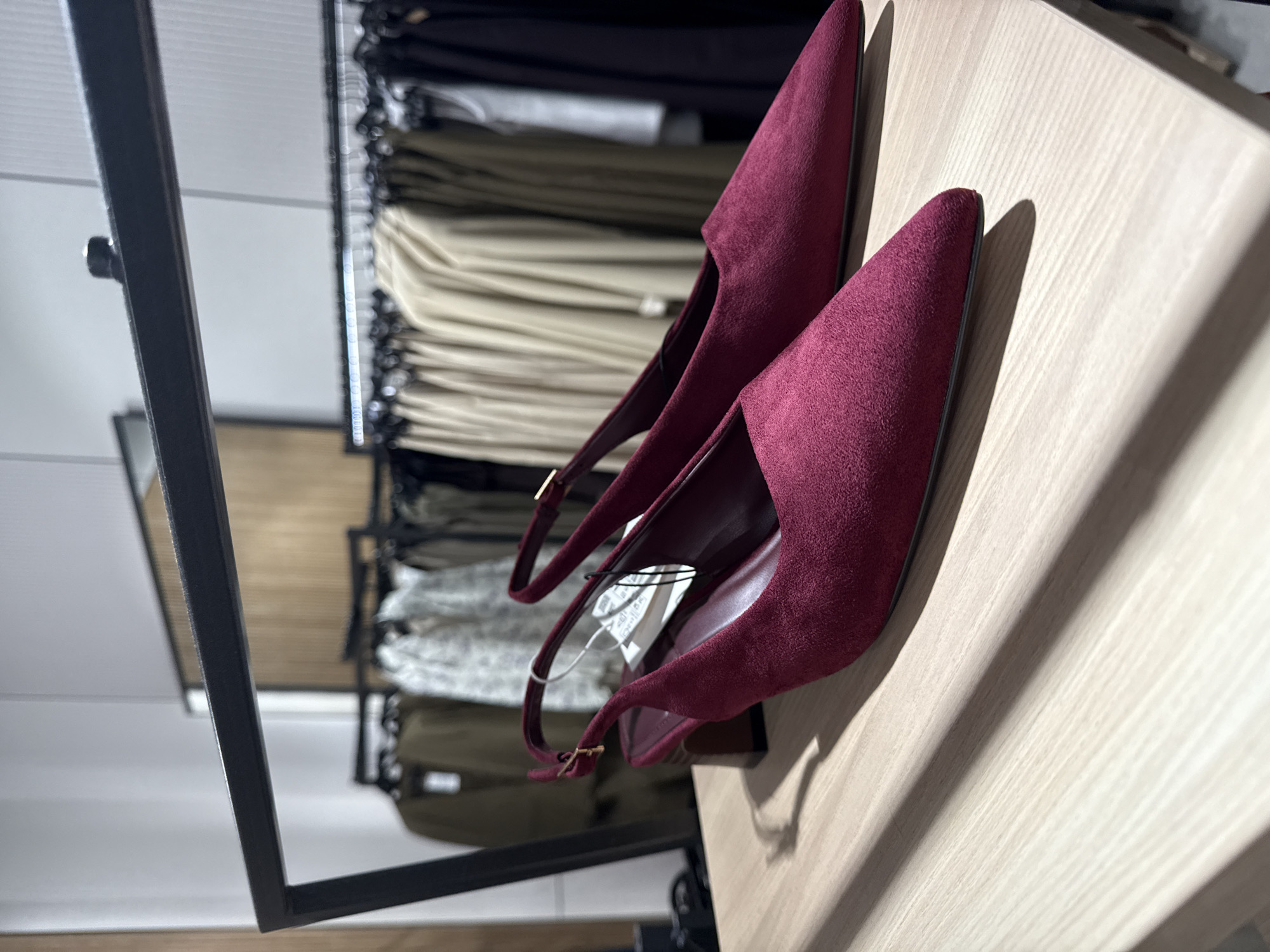This image captures a pair of deep maroon or burgundy suede slingback high heels, prominently displayed on a light-colored wooden surface, likely a shelf or table within a retail environment. The shoes feature pointed toes and a sleek design, with a small gold-toned buckle visible on the strap of one shoe.
Bright, possibly artificial, lighting illuminates the scene, casting distinct shadows from the shoes onto the wooden surface, which exhibits a subtle wood grain texture. In the background, out of focus, are racks of clothing. These garments appear to be various items like trousers and tops, neatly hung and predominantly in neutral colors such as beige, olive green, white, and dark shades. A black metal frame, part of the store fixtures, is visible on the left side and in the background.
A small white tag, likely a price or product information tag, hangs from one of the shoes, though its text is illegible. No people are visible, and the setting strongly suggests an indoor retail store, possibly in Varna, Bulgaria, where merchandise is presented for sale.