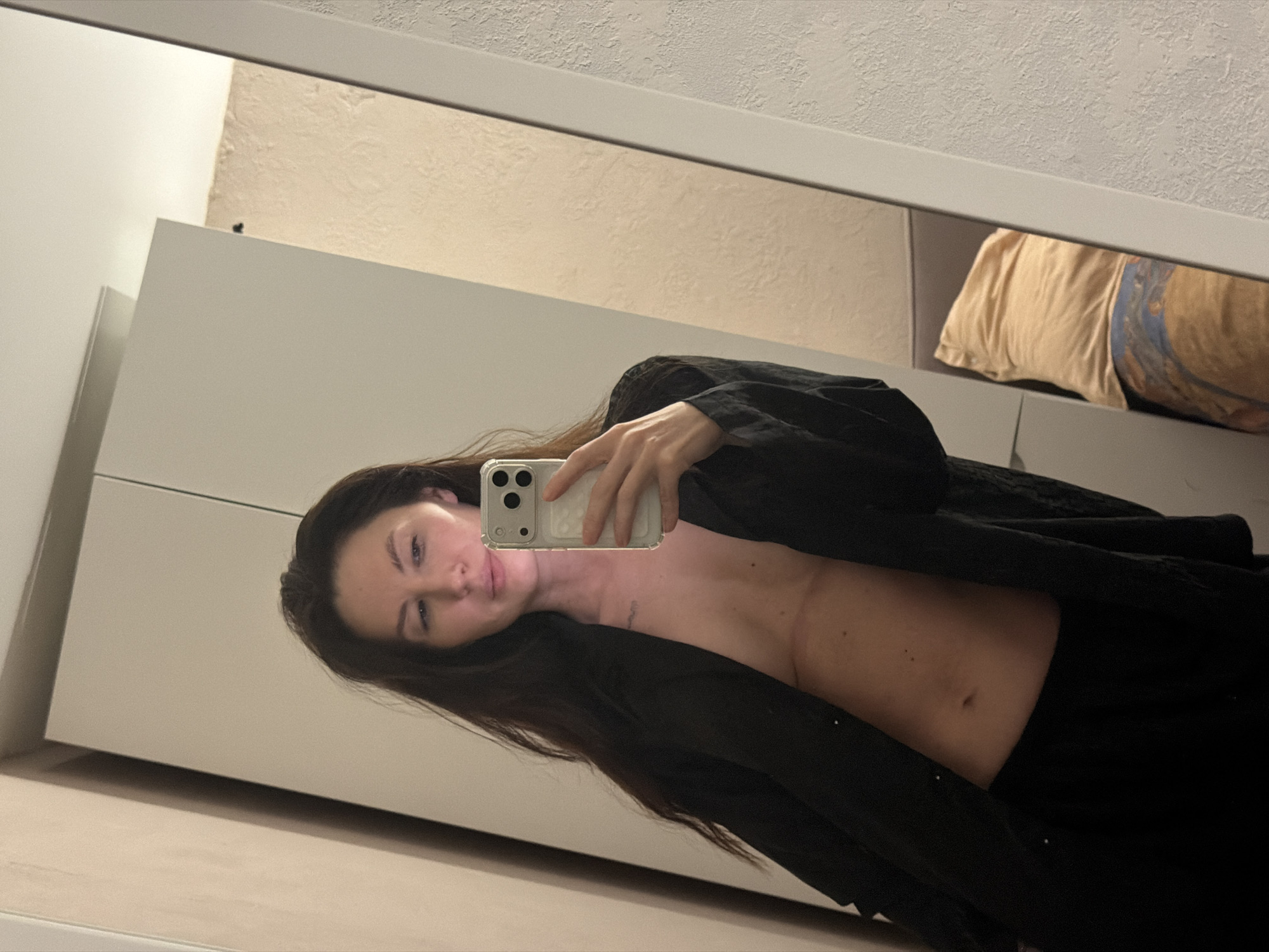 A mirror selfie taken indoors. The person is holding a phone in their right hand and angling it towards a mirror. The person has long, dark hair and is wearing a black outfit open at the chest. Visible in the background, reflected in the mirror, is a cream-colored pillow. The environment appears to be a residential room with a white dresser or cabinet. There is no information to indicate this photo was taken in the city of Perm, Russia.