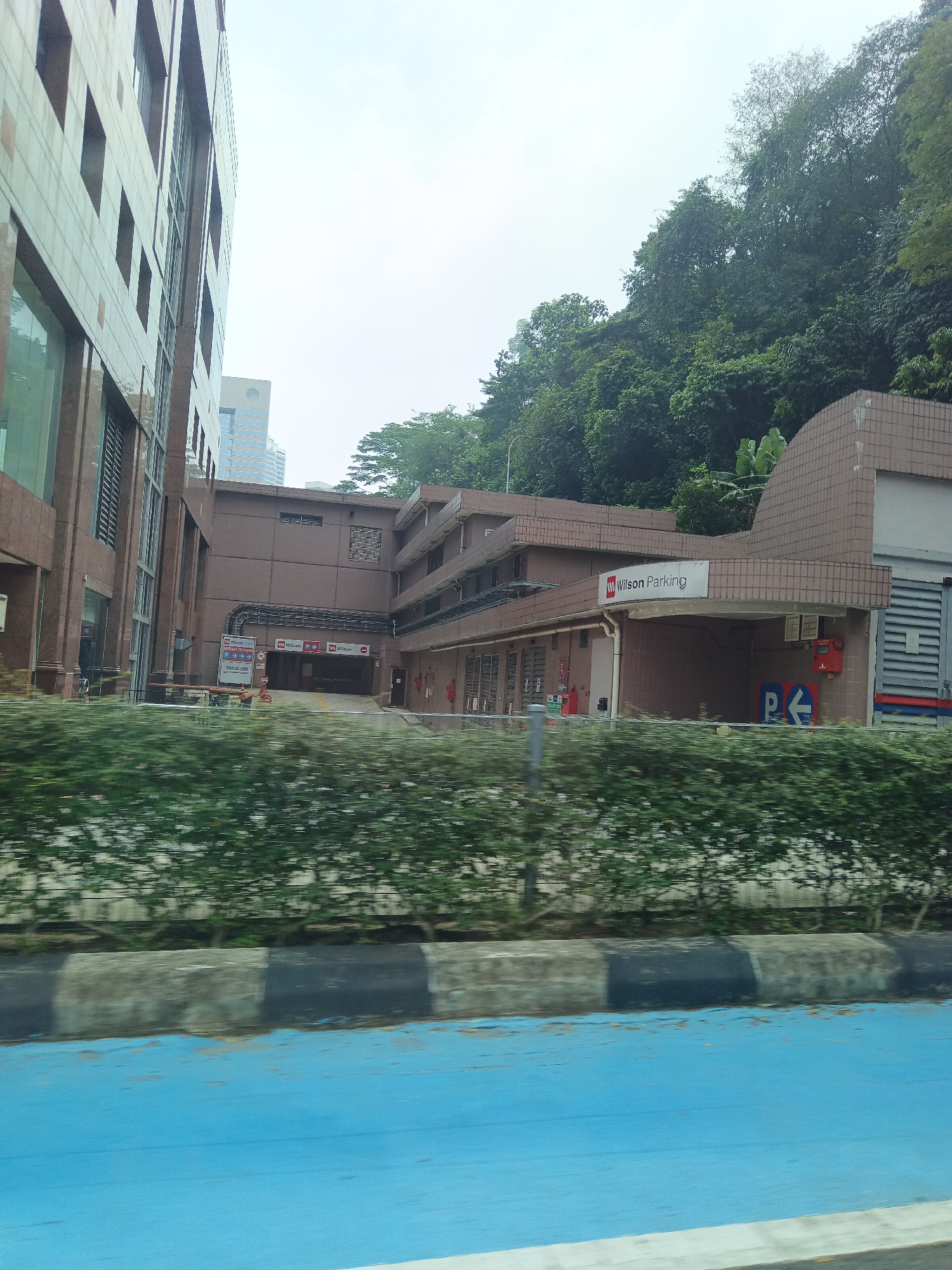 The image captures a view of a Wilson Parking facility in Kuala Lumpur, Malaysia, located at the base of a lush, tree-covered hill. The parking entrance is visible, with a sign displaying "Wilson Parking" prominently above it. To the left, a large, modern office building with a patterned facade stands tall, its windows reflecting the overcast sky. A dense hedge runs along the foreground, partially obscuring a blue-painted surface and a striped curb. The overall scene suggests an urban environment where modern infrastructure meets natural landscape.