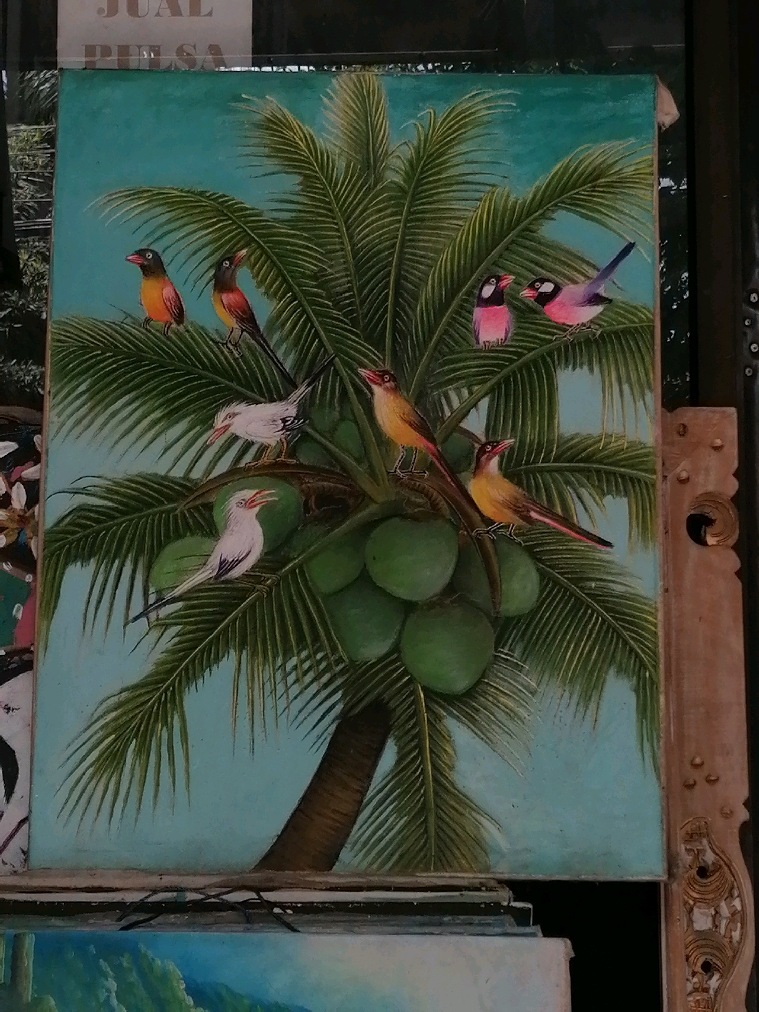 The image shows a painting of a coconut tree with green coconuts and several colorful birds perched on its fronds and branches. The sky is a light teal color. There is text in the background, partially obscured, that appears to say "JUAL PULSA". The painting is framed by an ornate wooden carving on the right side. The painting is propped up against other items, suggesting it is displayed for sale in an outdoor market or shop. The scene is likely located in the city of Ubud District, Indonesia, known for its vibrant art scene and traditional markets.