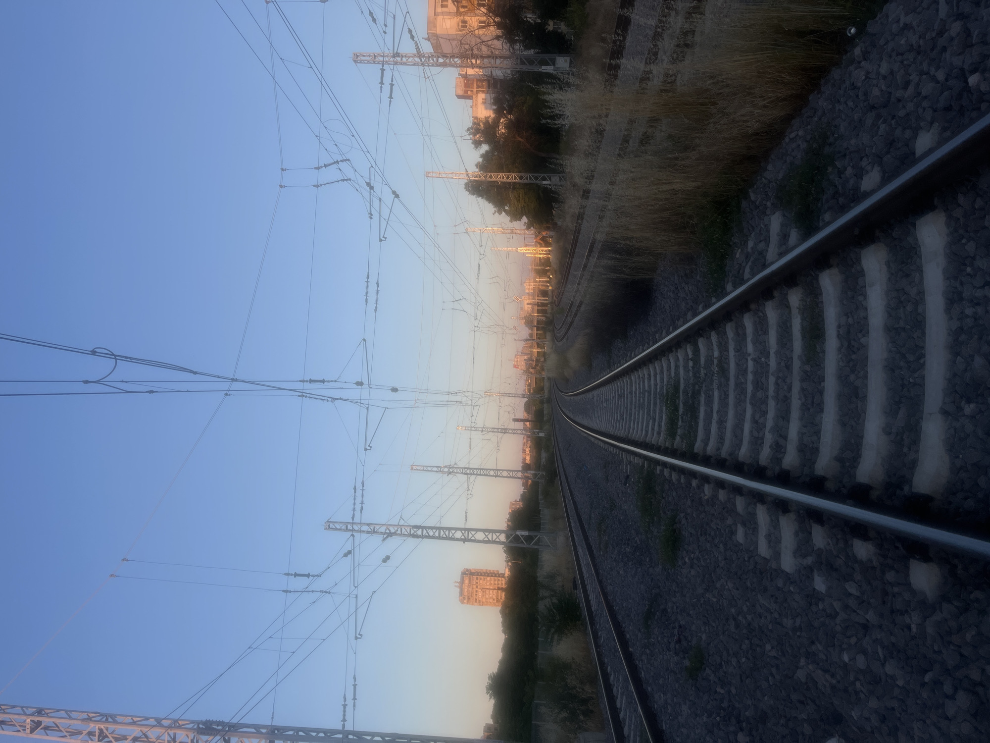The image depicts a train track curving to the right in the foreground. On either side of the tracks are gravel beds and dry vegetation. Overhead, a complex network of electrical wires and support poles stretches across the scene, indicating an electrified railway. In the background, several buildings are visible, including a tall, multi-story building to the left and other structures to the right. The sky is clear and blue, transitioning to a lighter hue near the horizon, suggesting it is either early morning or late evening. Two figures are visible in the far distance, standing on what appear to be the overhead wires, with one figure in an orange safety vest. The scene is an urban or semi-urban environment with infrastructure for train transportation.