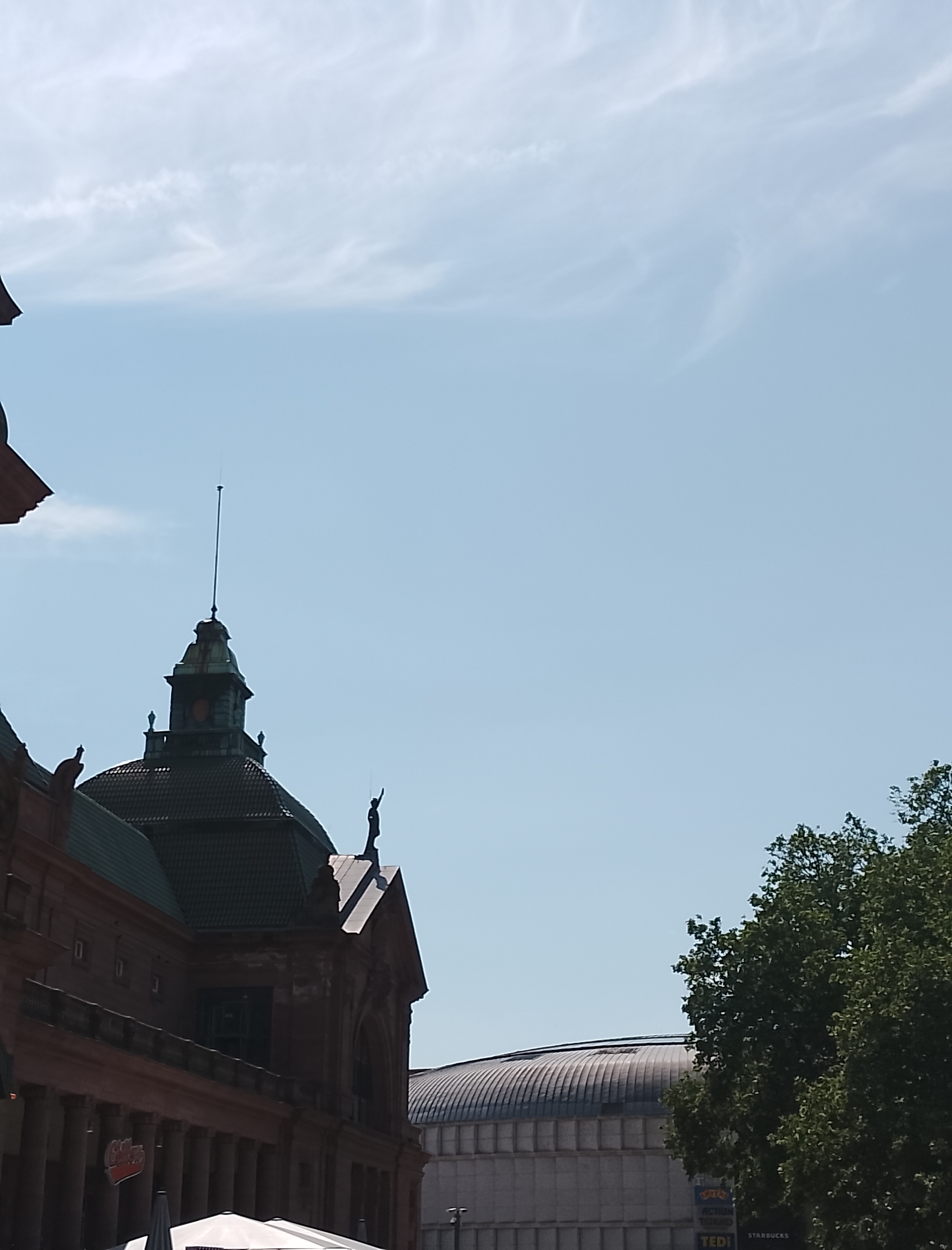 The image shows the upper portion of an ornate brick building with a domed roof and a tall spire. A silhouette of a person stands on the peak of a smaller dome to the right of the main spire. The building is partially obscured by a large green tree on the right side of the frame. In the background, a large, modern building with a curved roof is visible. The sky is a clear blue with a few wispy clouds. The lighting suggests it is daytime, likely late morning or early afternoon, with bright sunlight.
Visible text includes a red sign on the building that appears to say "Glocken" or a similar word with multiple letters, and a sign below it that says "STARBUCKS". Other signs in the background are too small to read clearly.
The scene depicts an urban environment with historical architecture juxtaposed with modern structures and lush greenery. There are no active events clearly depicted, and the overall mood is serene and observational.