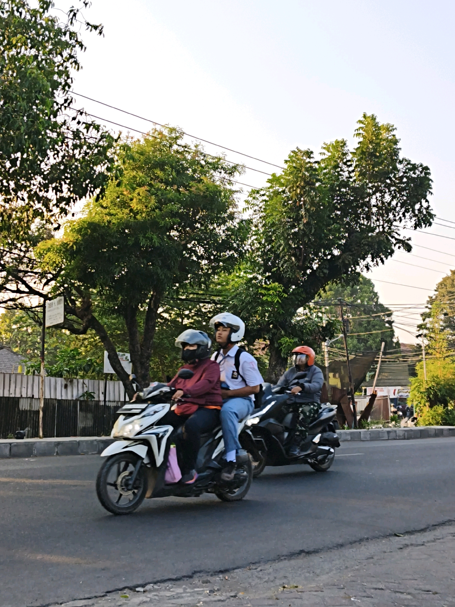 In the city of Surakarta, Indonesia, three individuals are traveling on two motorcycles on a paved road. The foreground shows a white scooter with two occupants. The front rider wears a maroon jacket and a grey helmet with a face mask. The passenger behind them is wearing a white school uniform with a blue tie and a white helmet. A pink bag is visible hanging from the front of the scooter. Behind them, a second, darker-colored motorcycle is ridden by a person wearing a grey jacket and an orange helmet. They are wearing camouflage-patterned pants. The background features lush green trees, a wooden fence, and overhead power lines. The lighting suggests it is either early morning or late afternoon, with warm sunlight filtering through the trees.