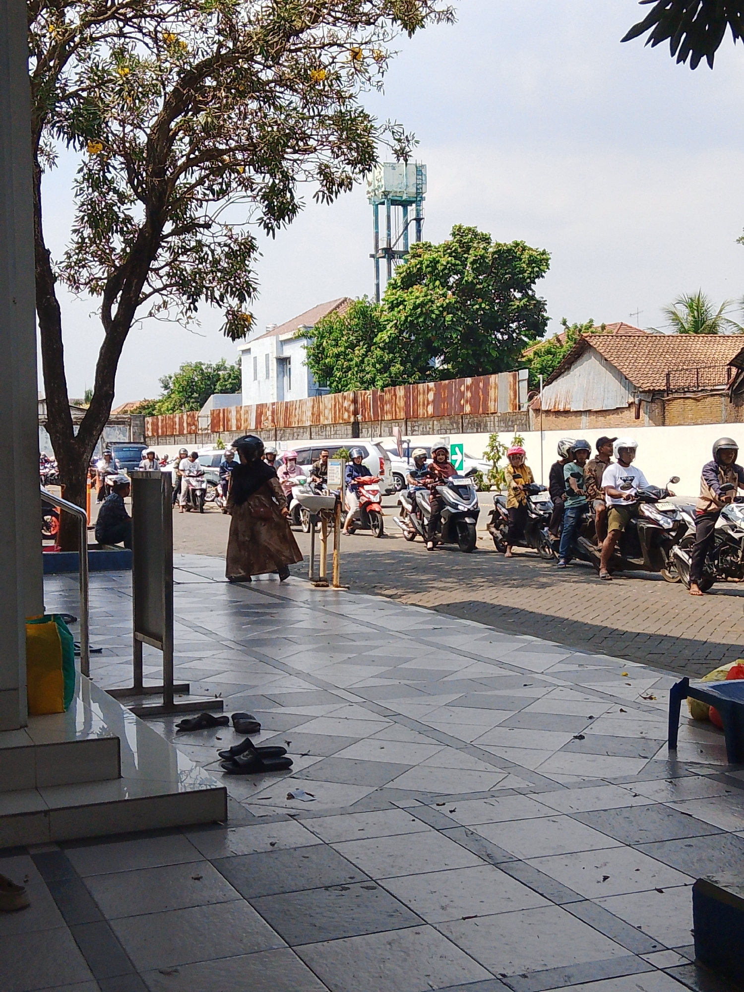 This is a vertical, eye-level, medium shot taken on a sunny day in the city of Potroyudan, Indonesia, showing a busy street scene. In the foreground, a tiled walkway features several pairs of black flip-flops. A large tree with green leaves and yellow flowers dominates the left side of the frame, its trunk partially obscuring a white wall and a blue and yellow bag. Beyond the walkway, a street is filled with people on motorcycles, forming a queue. Most individuals are wearing helmets. A woman in a long, patterned dress and a black hijab is walking across the street, while other pedestrians and motorcyclists are visible in the background. Buildings with tiled roofs line the street, and a tall water tower stands in the distance against a bright sky. A rusty metal fence runs along the edge of the street.