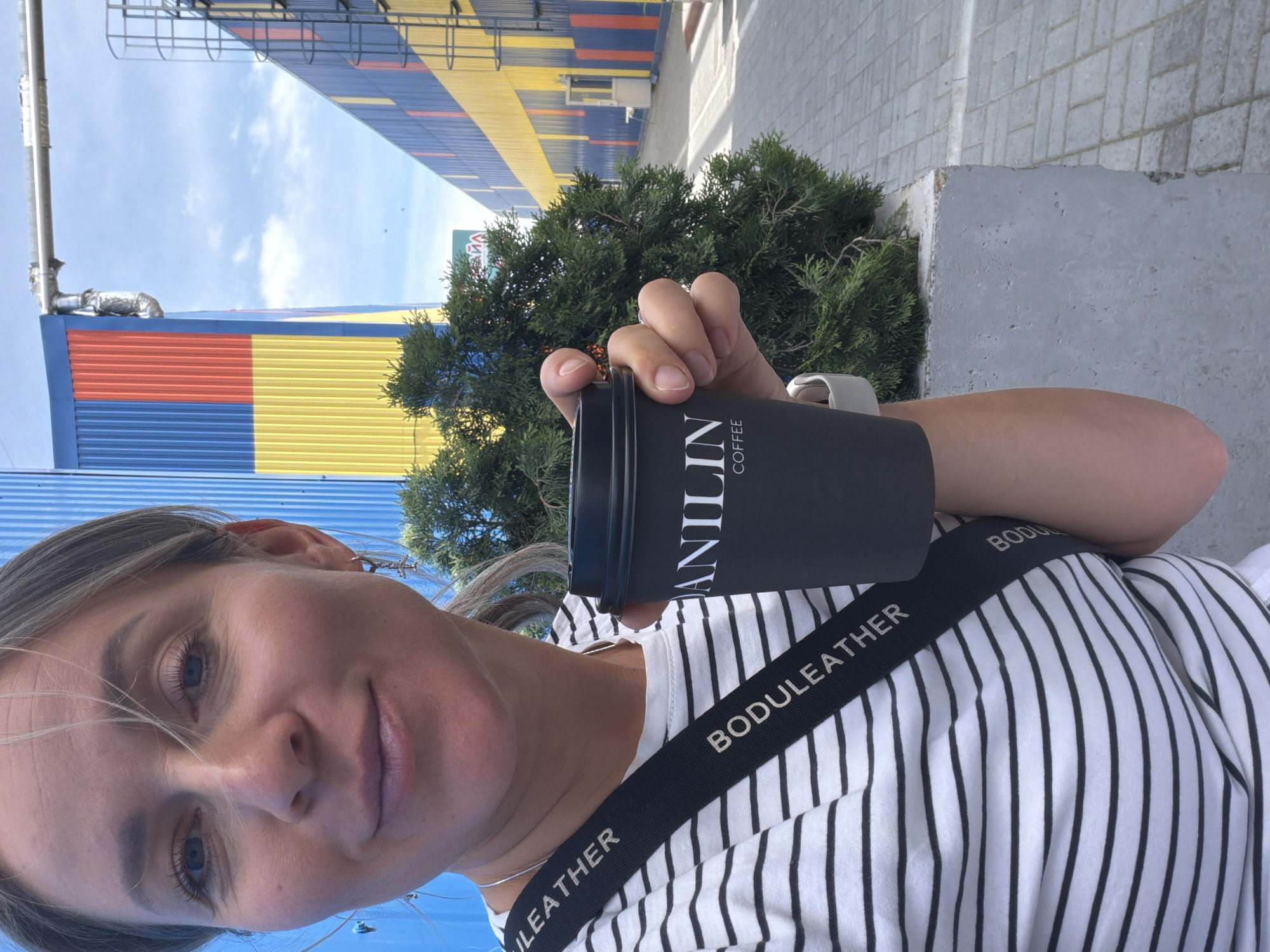 A person is holding a "VANILIN COFFEE" mug in their right hand. They are wearing a striped shirt and a black strap labeled "BODULEATHER". Behind them is a green bush and a building with blue, red, and yellow panels. The location is Chaikovsky, Russia.