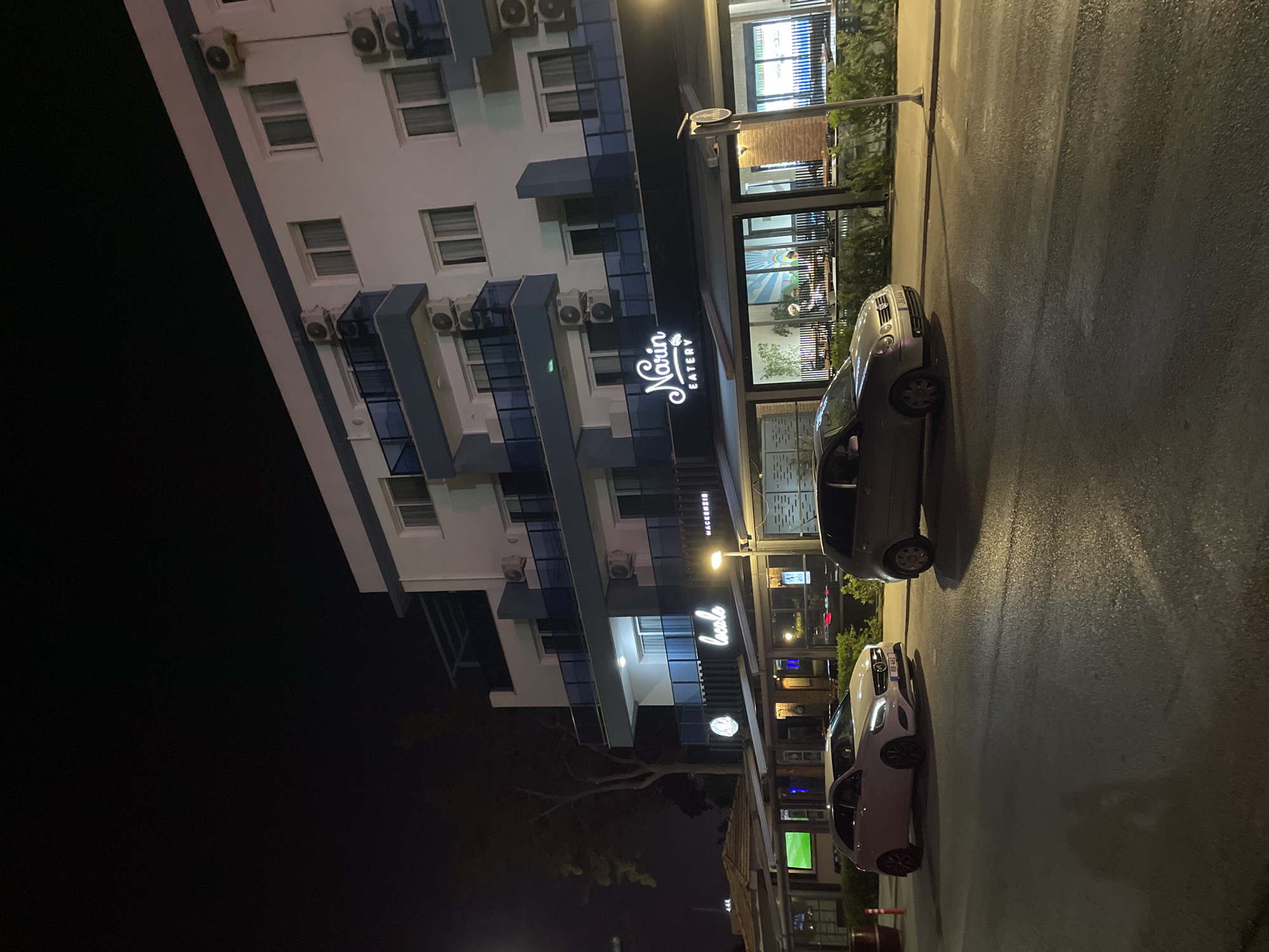 The image captures a nighttime street scene in Trikomo, Northern Cyprus. A multi-story building with white walls and blue accents dominates the background. The building features multiple windows and air conditioning units mounted on its exterior. In front of the building, a row of businesses with illuminated signs is visible. One sign reads "Narin EATERY" in stylized white lettering. Another sign displays "locale" in similar font. A third sign appears to be for "MACKENZIE". The businesses have large glass fronts, revealing indoor seating areas with tables and chairs.
Two cars are parked on the wet asphalt road in the foreground. The car on the left is a white Mercedes-Benz, and the car on the right is a silver Volkswagen. A pedestrian crossing is marked on the road. Streetlights and interior lighting from the businesses illuminate the scene. A tree with sparse foliage stands to the left of the buildings, partially obscuring the view. The overall atmosphere is that of a commercial area at night.